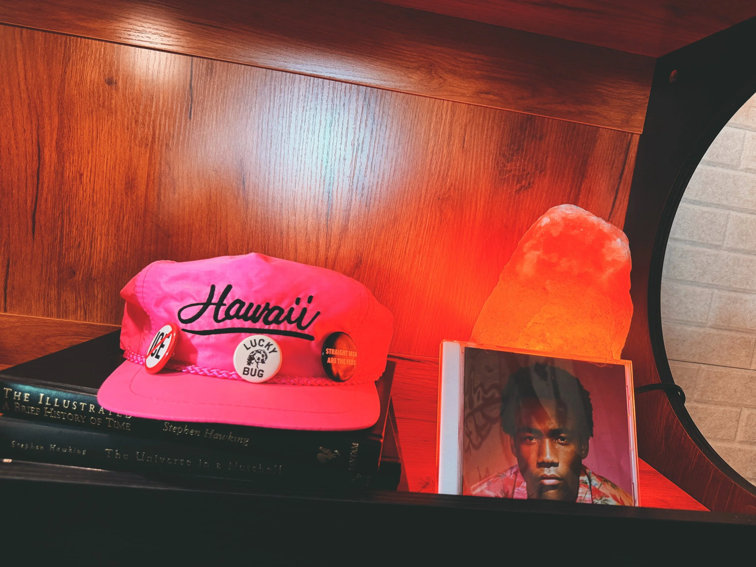 A pink hat with the word "Hawaii" and various pins, including a red button that says "Straight Men Are the Feds", atop a stack of books, next to a Himalayan salt lamp, on a black surface against a wooden background.