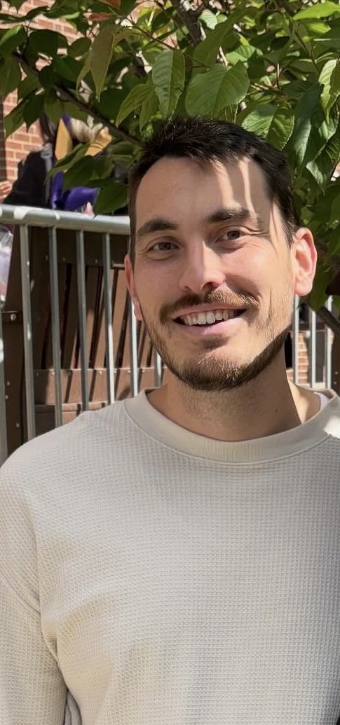 A smiling man with dark hair and a beard outdoors, standing in front of green leafy plants and a brick building.