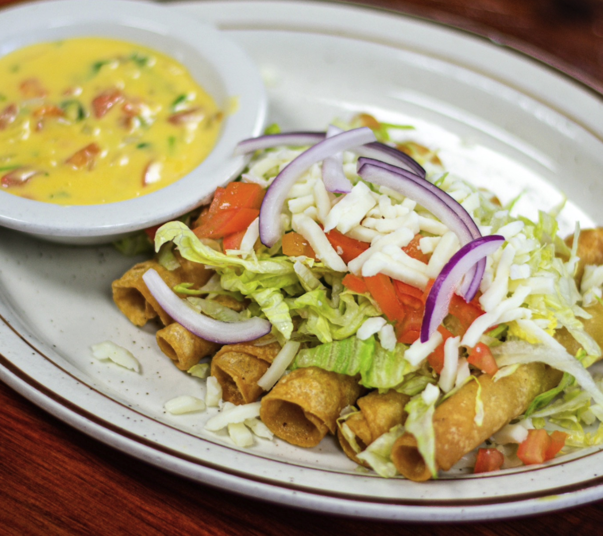 Fried taquitos topped with shredded cheese, lettuce, tomatoes, and red onion, served with a side of creamy yellow sauce with chopped peppers.