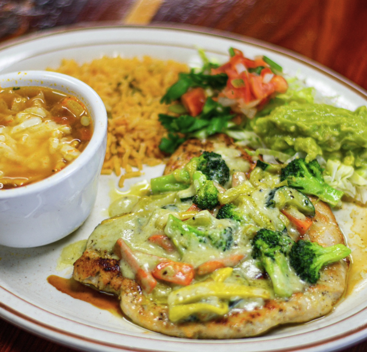 Plate of food with chicken topped with broccoli and mixed vegetables, a side of guacamole, pico de gallo, rice, and a bowl of cheese dip.