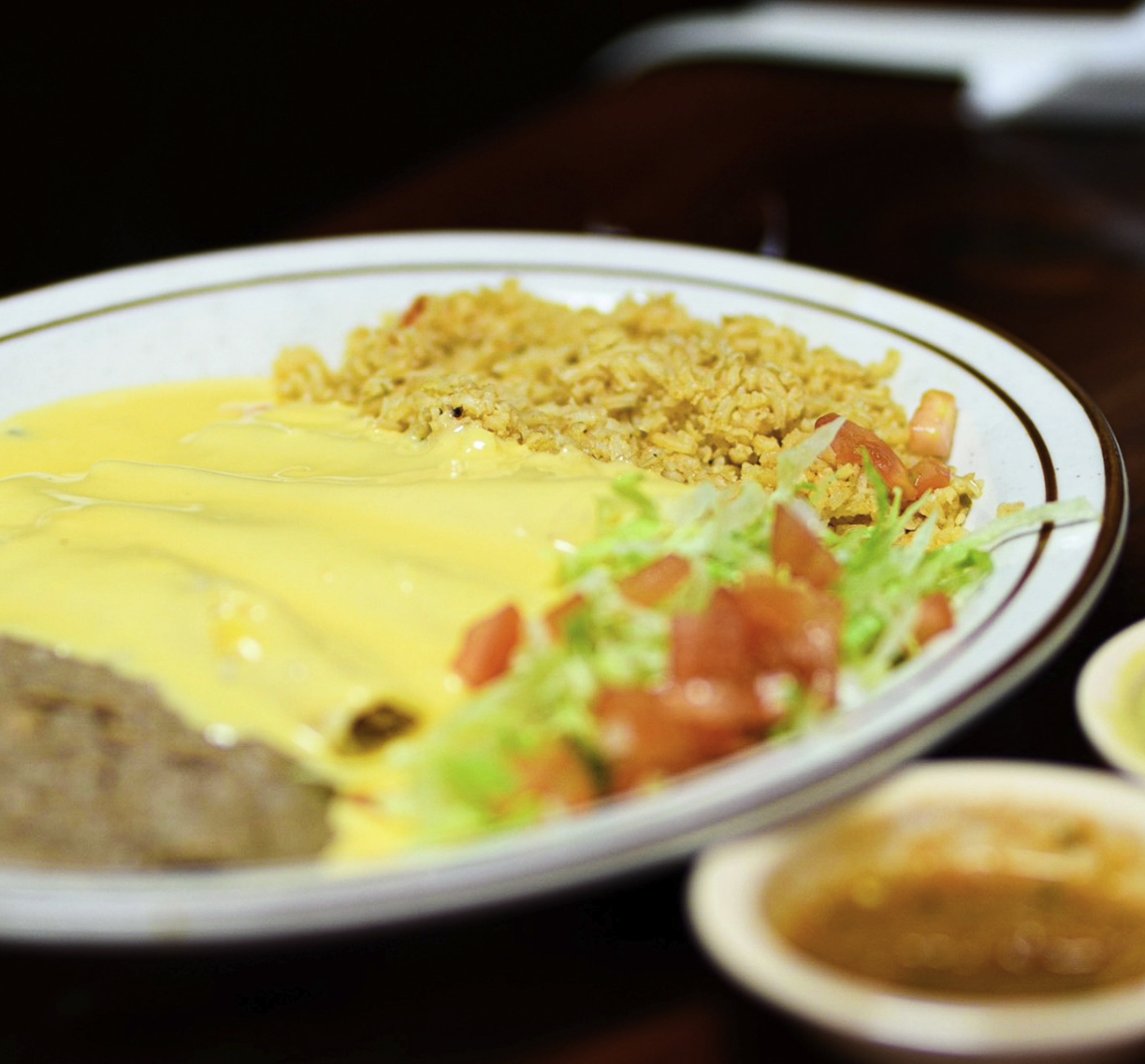 A plate with rice, scrambled eggs, shredded lettuce with diced tomatoes, and a beef taco with cheese, all on a dark table. A side dish of sauce is partially visible.