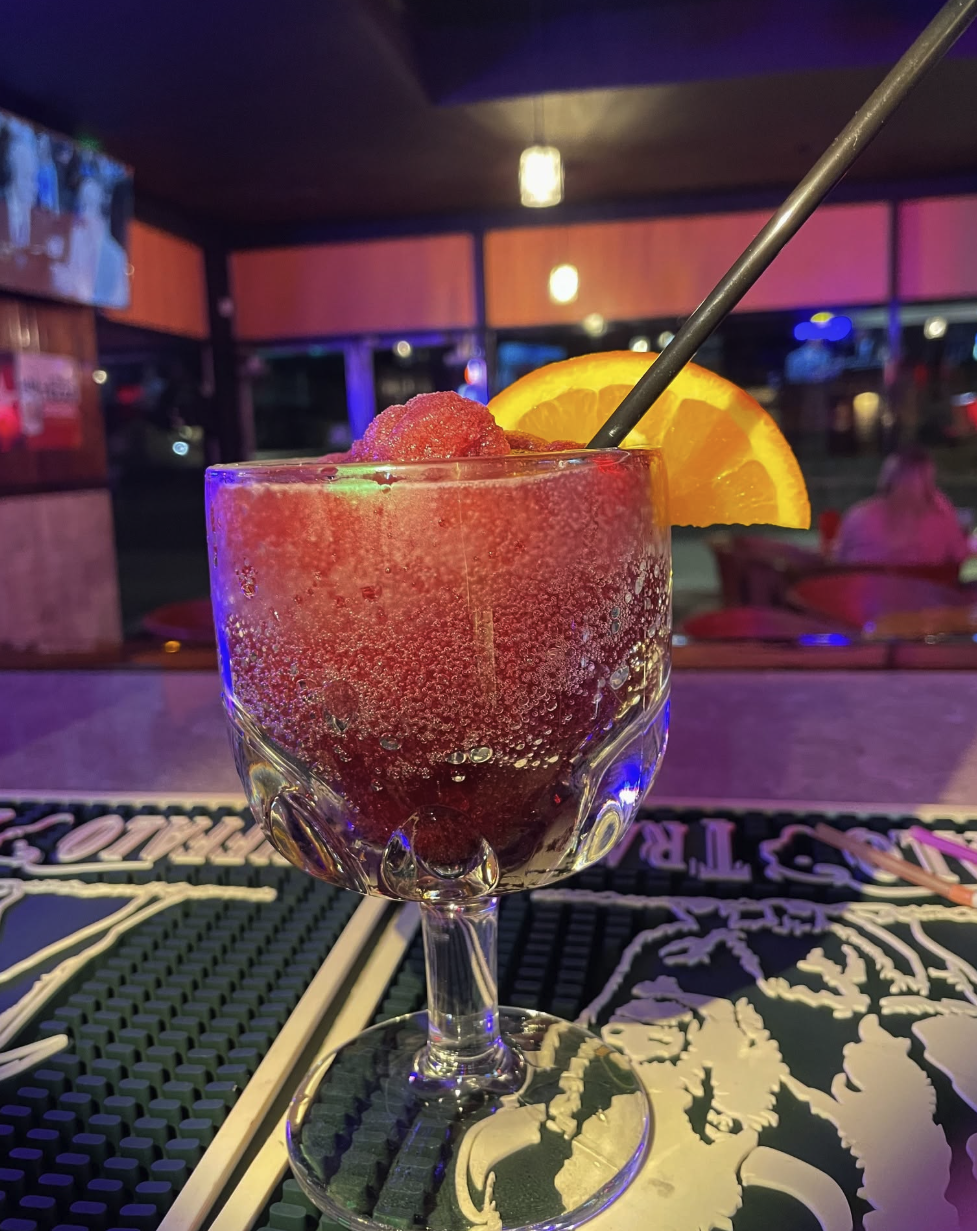 A glass of red frozen cocktail garnished with an orange slice and a black straw, on a bar counter with a colorful dimly lit background.