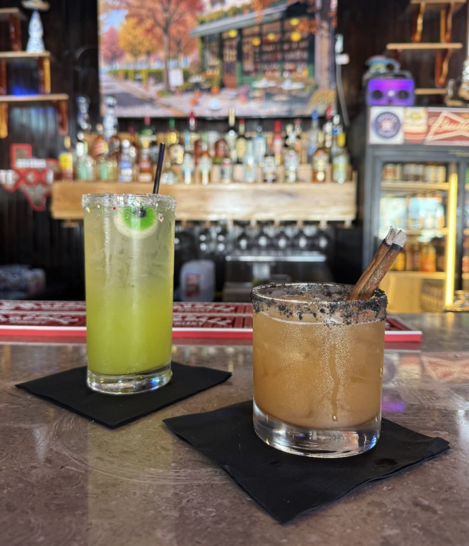Two cocktails on a bar counter, one green and one amber, with a bar and liquor bottles in the background.