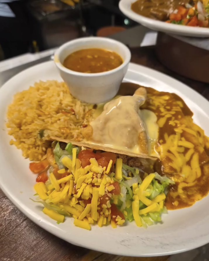 Plate of Mexican food with rice, salad, chicken covered in cheese sauce, refried beans, and a small bowl of sauce.