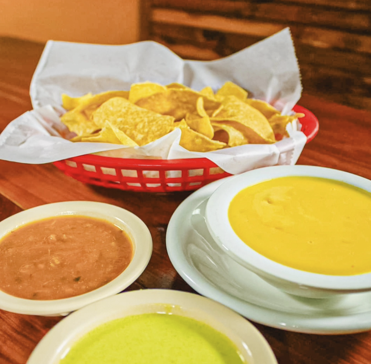 Basket of tortilla chips with three bowls of salsa and nacho cheese dip on a wooden table.