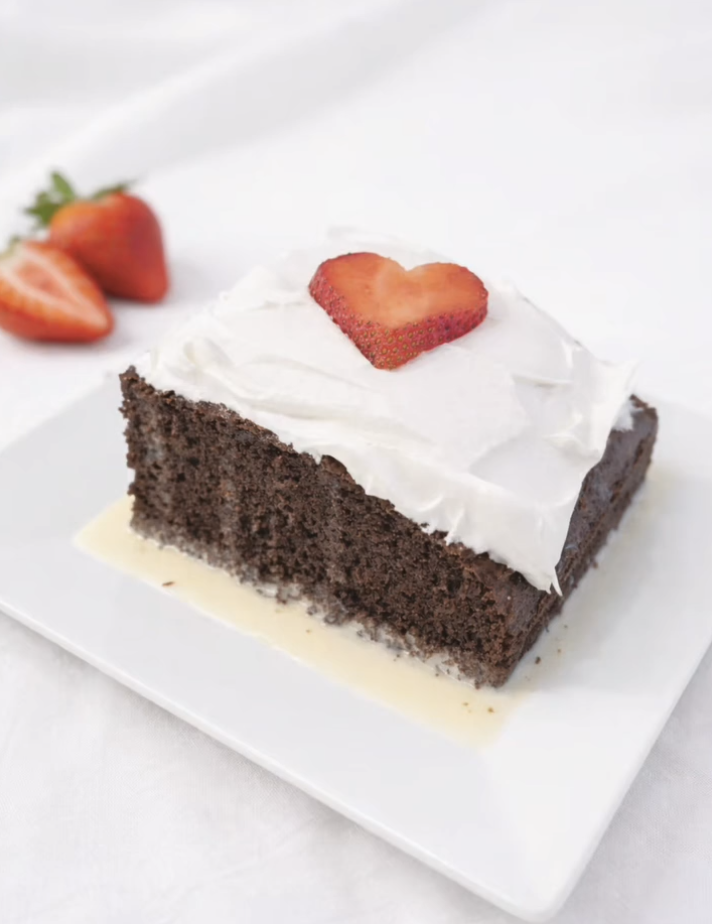 Chocolate cake with white frosting and a sliced strawberry on top, served on a white plate, with whole strawberries in the background.