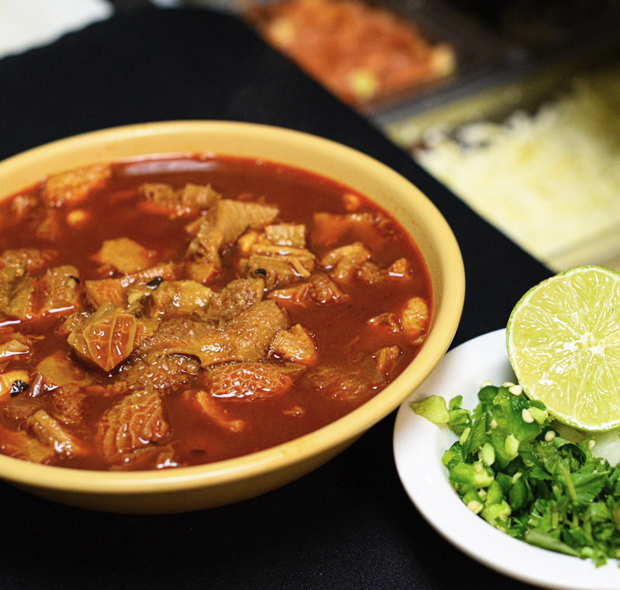 Bowl of beef stew with carrots and potatoes, garnished with lime and chopped green onions, with side dishes of sauce and spicy chili in the background.