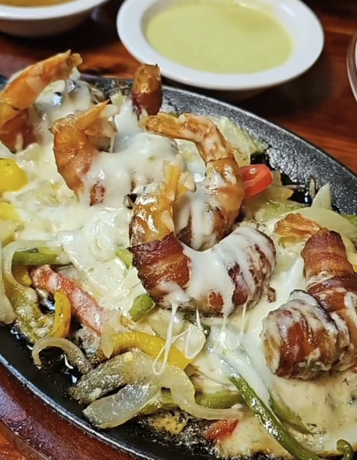 Shrimp wrapped in bacon served on a sizzling plate with melted cheese, accompanied by sliced onions and green and yellow bell peppers, with a bowl of creamy sauce in the background.