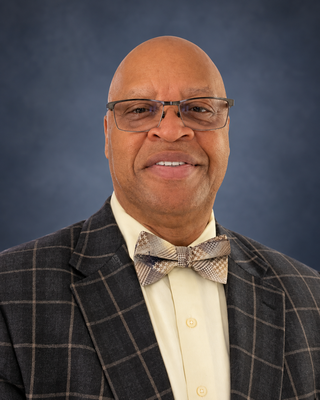 A middle-aged man with a bald head and glasses, wearing a plaid blazer, a light yellow shirt, and a patterned bow tie, smiling at the camera. Background is dark blue.