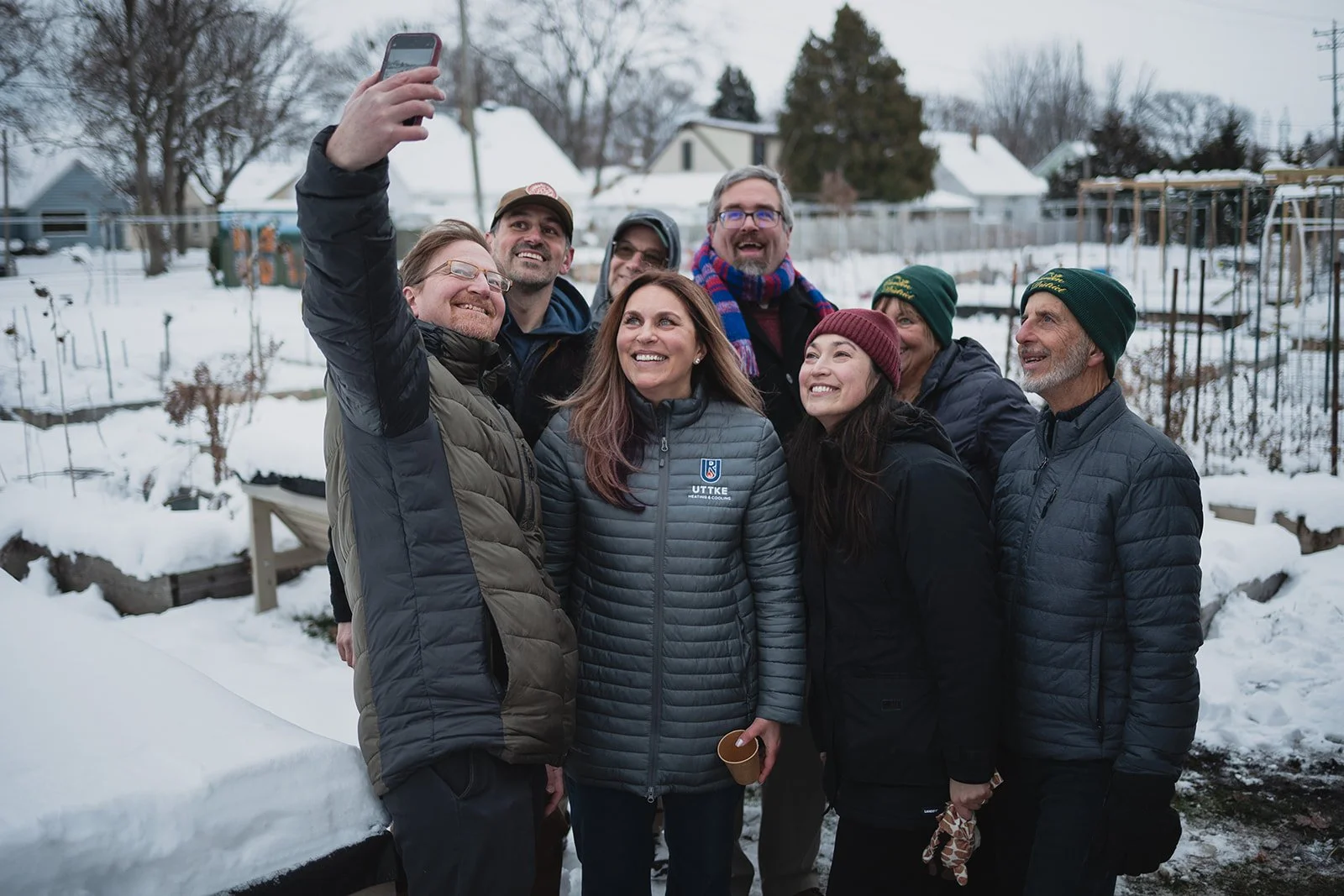 A group of eight people taking a selfie outdoors in snowy weather, dressed in winter jackets and hats, smiling and standing close together.