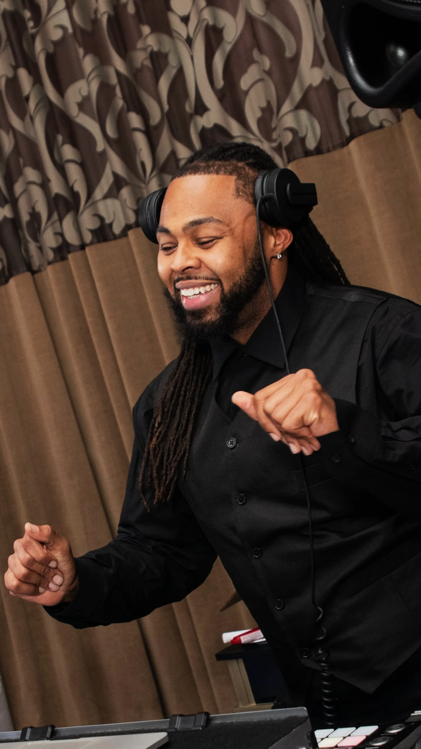 A man with long dreadlocks, a beard, and wearing a black shirt and headphones, appears to be dancing or enjoying music in a room with brown curtains.