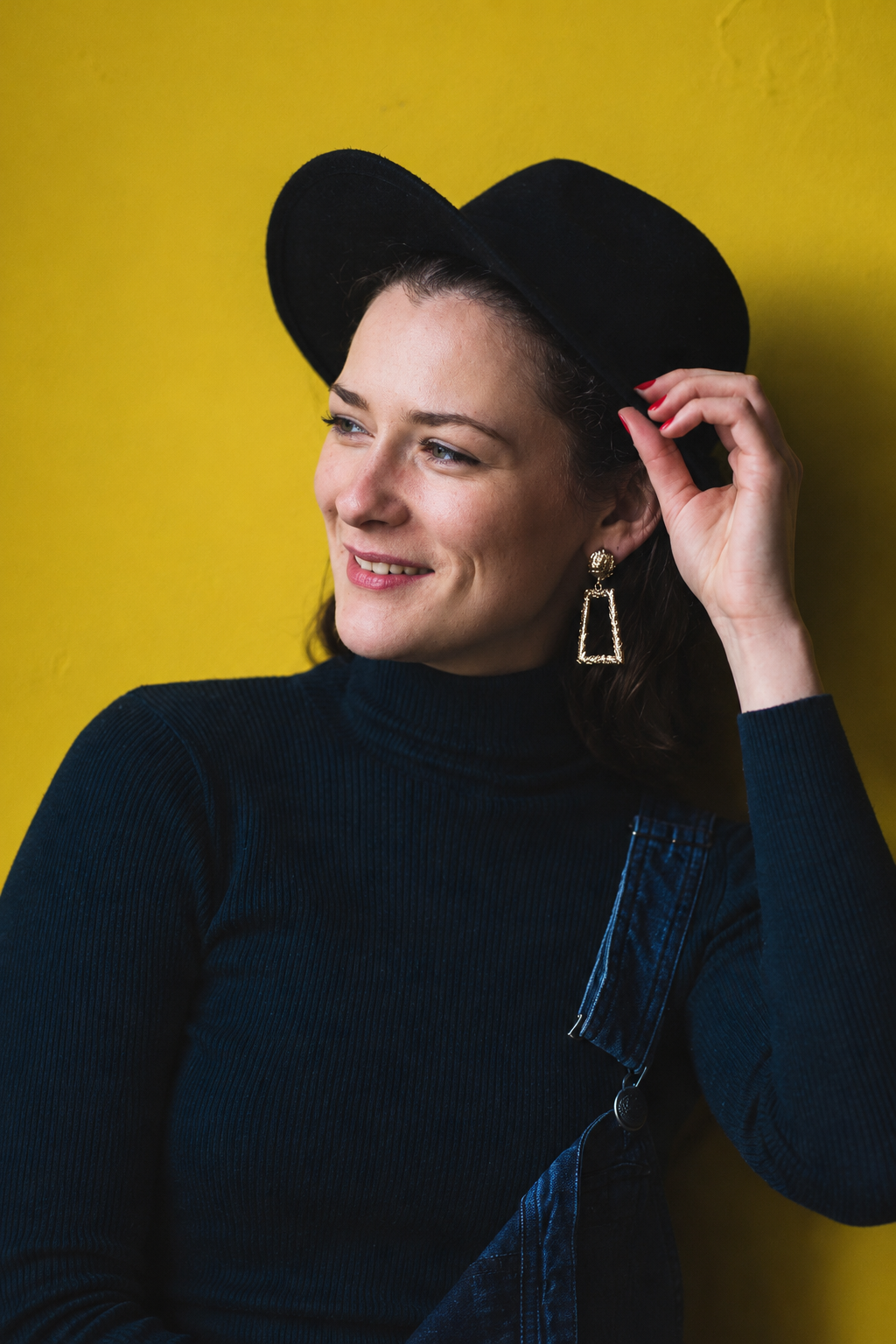 A woman wearing a black hat, navy blue turtleneck, and large gold earrings, smiling against a yellow background.