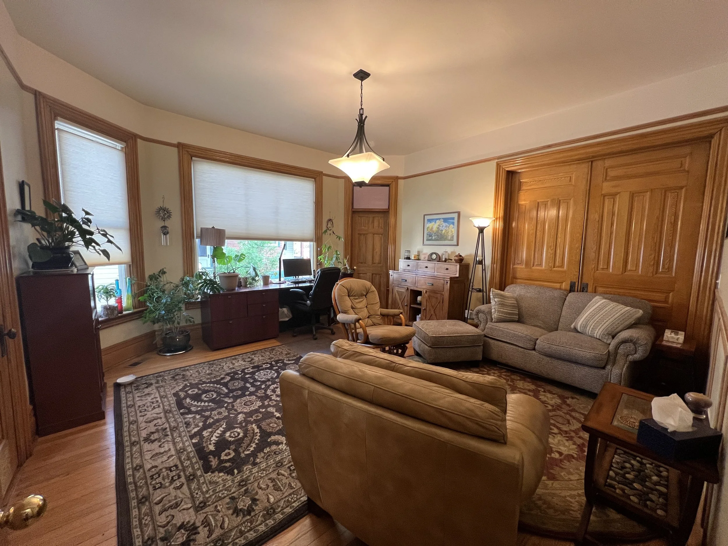 Living room with large windows, plants, and wooden furniture. Includes a sofa, armchairs, coffee table, sideboard, and decorative lamp, with a patterned area rug on a hardwood floor.