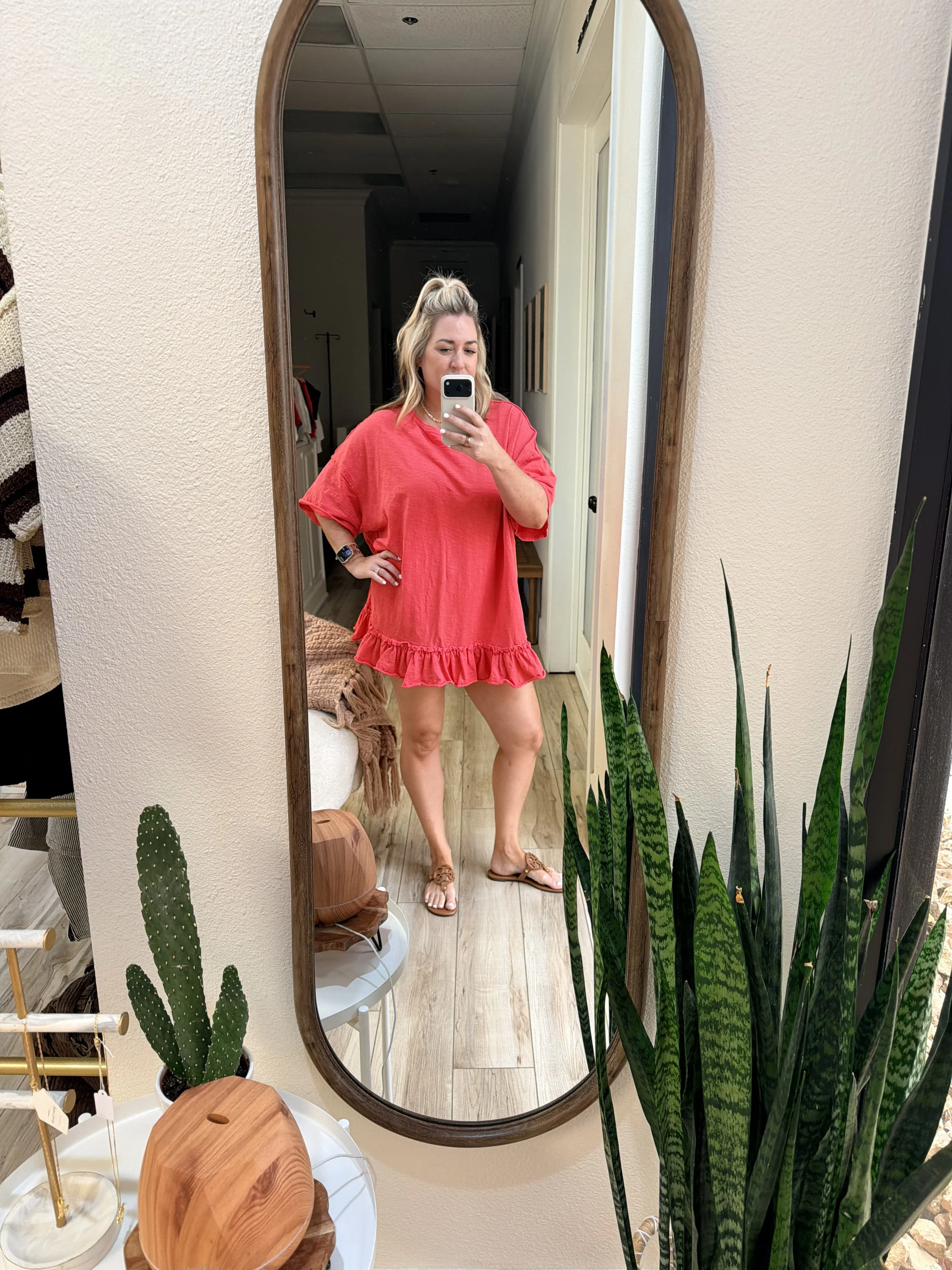 A woman taking a mirror selfie in a room with plants, wearing a red dress, sandals, and a smartwatch.