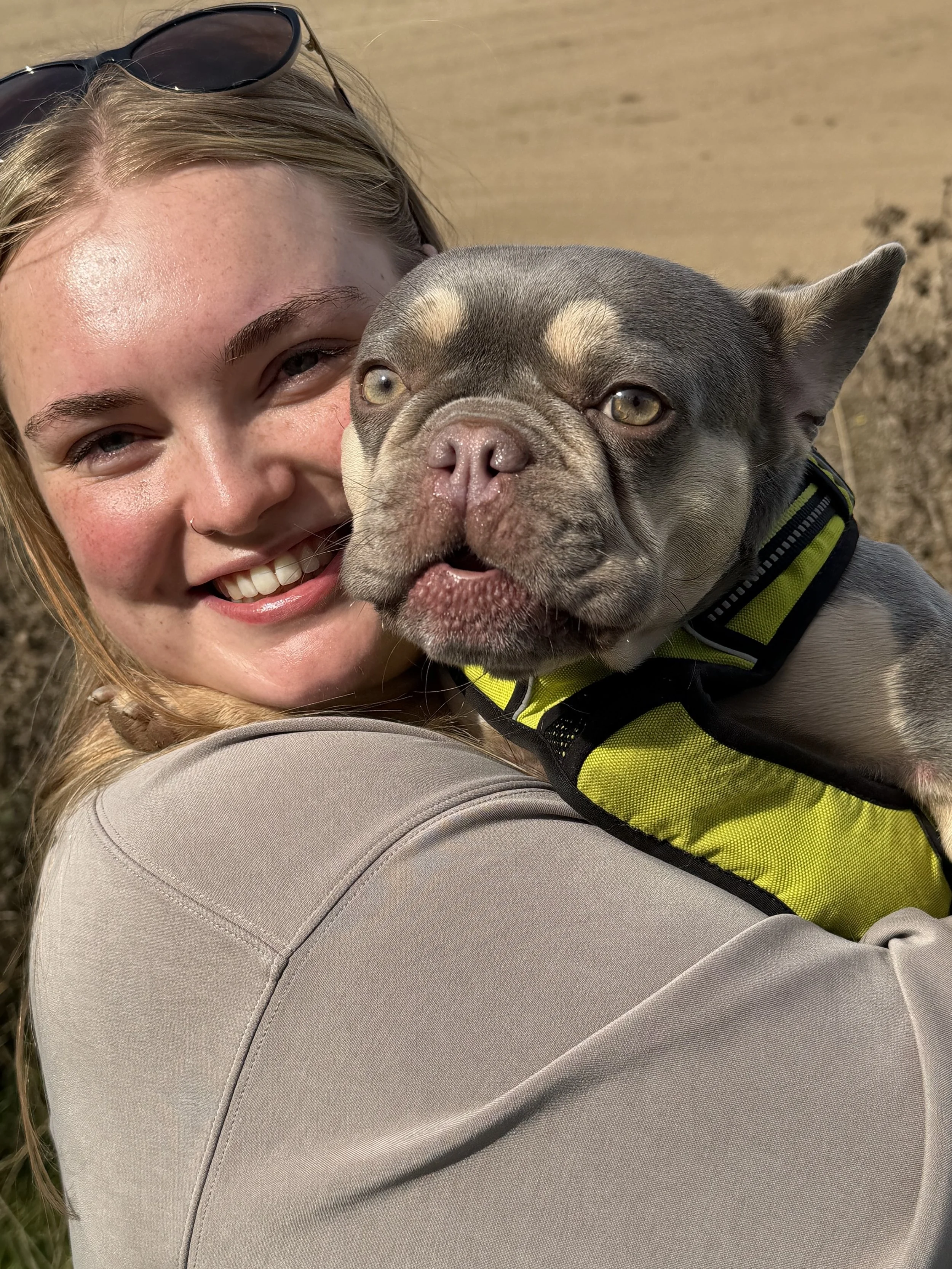 A woman with blonde hair wearing sunglasses on her head and a light-colored jacket, smiling while holding a gray and tan pitbull dog in an outdoor setting.