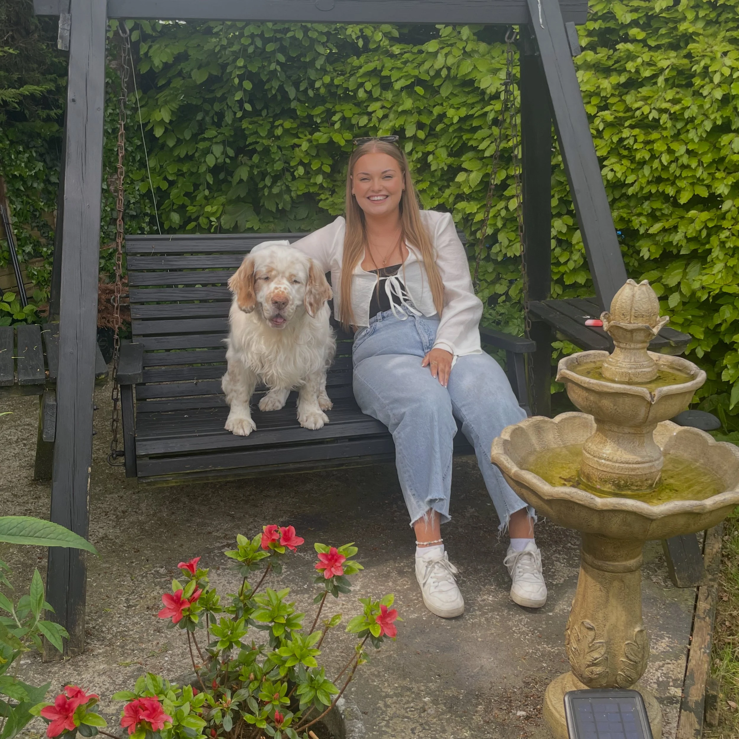 A young woman with long blonde hair sitting on a black wooden swing bench in a garden, smiling, with a white and brown dog sitting beside her. There are green leafy bushes in the background, pink flowers in the foreground, and a decorative stone fountain nearby.