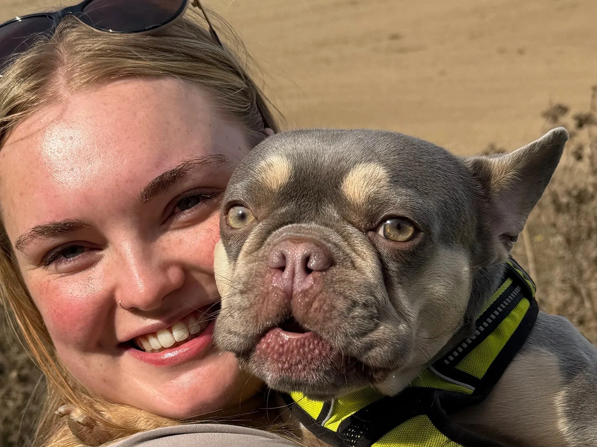 A woman with blonde hair and sunglasses on her head smiling with a dog, possibly a French Bulldog, with a brindle coat, with their faces close together, both looking at the camera.