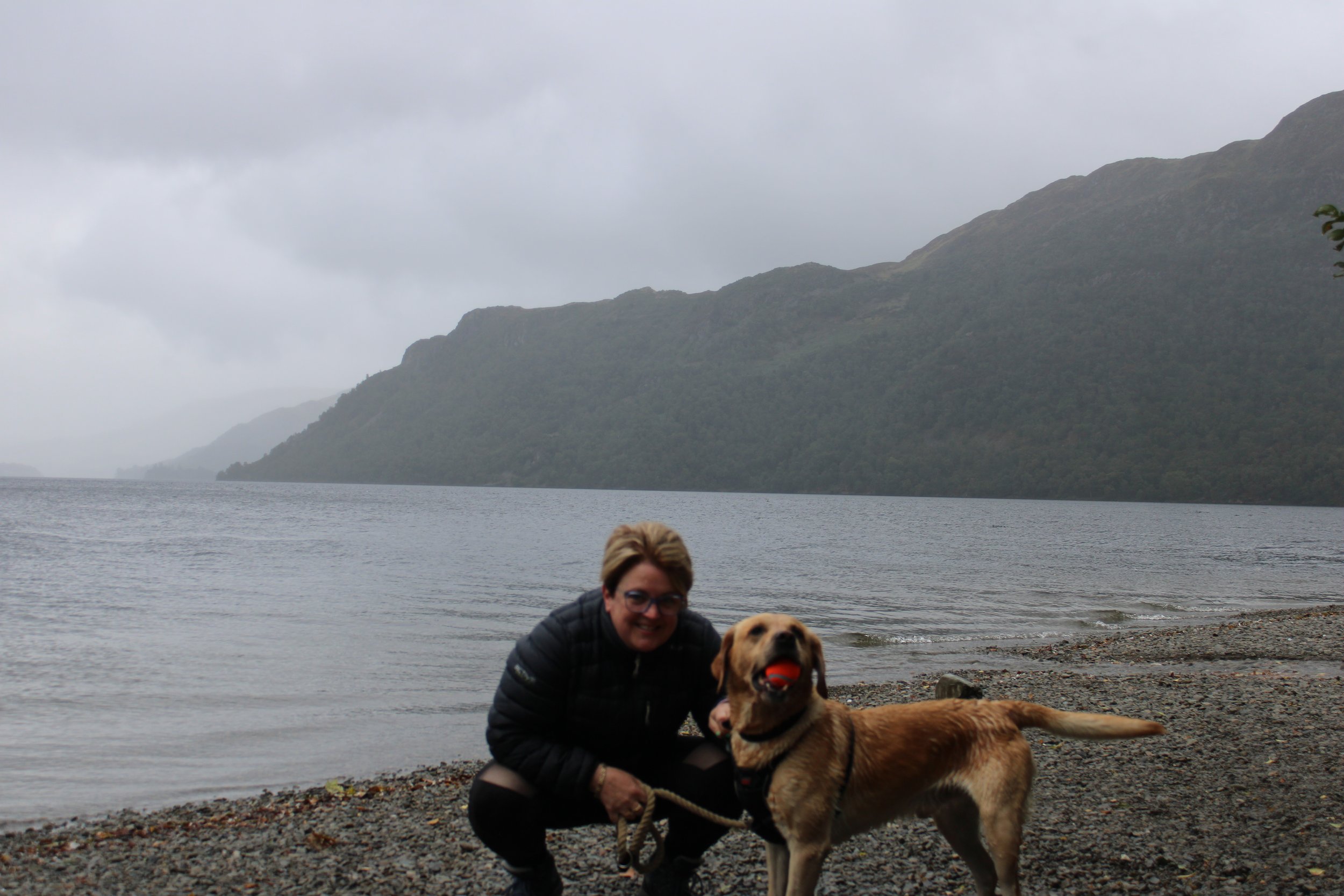 A woman and her dog at a lakeshore with mountains in the background under a cloudy sky.