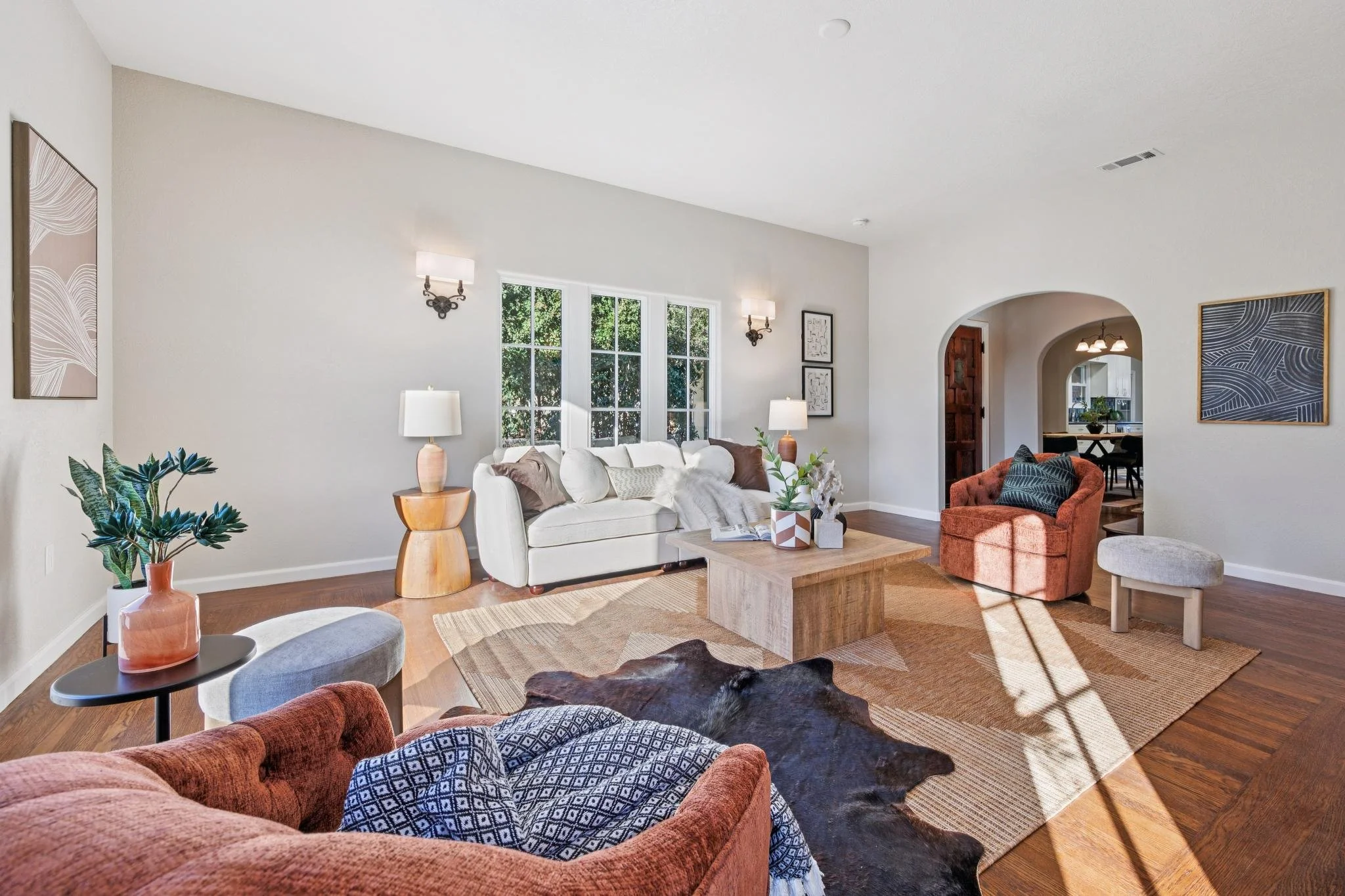 Living room with white walls, a white sofa with pillows, a glass top coffee table, an orange armchair, a black side table with a pink vase, artwork on the walls, a window with green trees outside, and hardwood floors.
