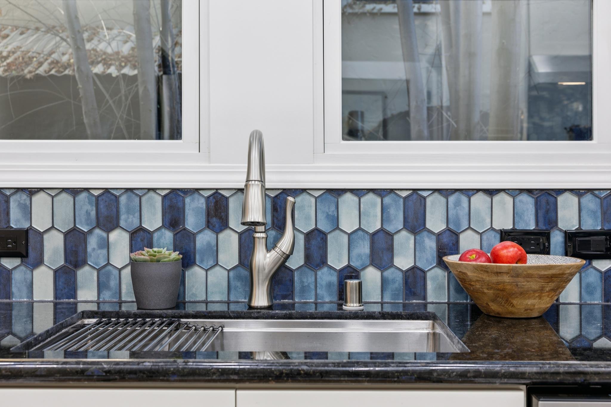Kitchen sink with a modern stainless steel faucet, a potted succulent plant on the left, a wooden bowl with red apples on the right, a blue hexagonal tile backsplash, and a window with a white frame above the sink.