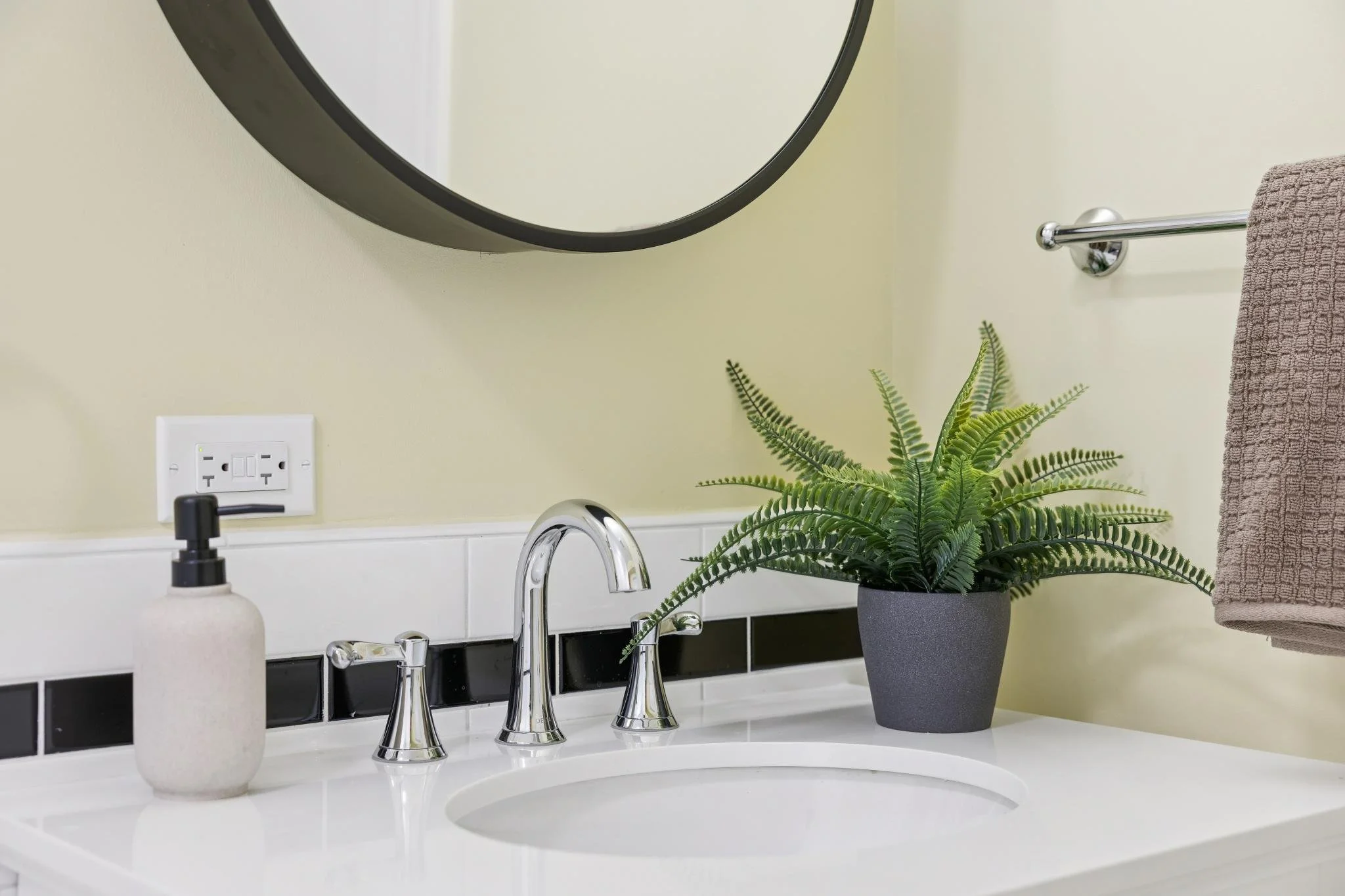 A bathroom sink area with a silver faucet, a soap dispenser, a potted green plant, a brown towel hanging on a towel rod, a mirror, a wall socket, and a black and white tiled backsplash.