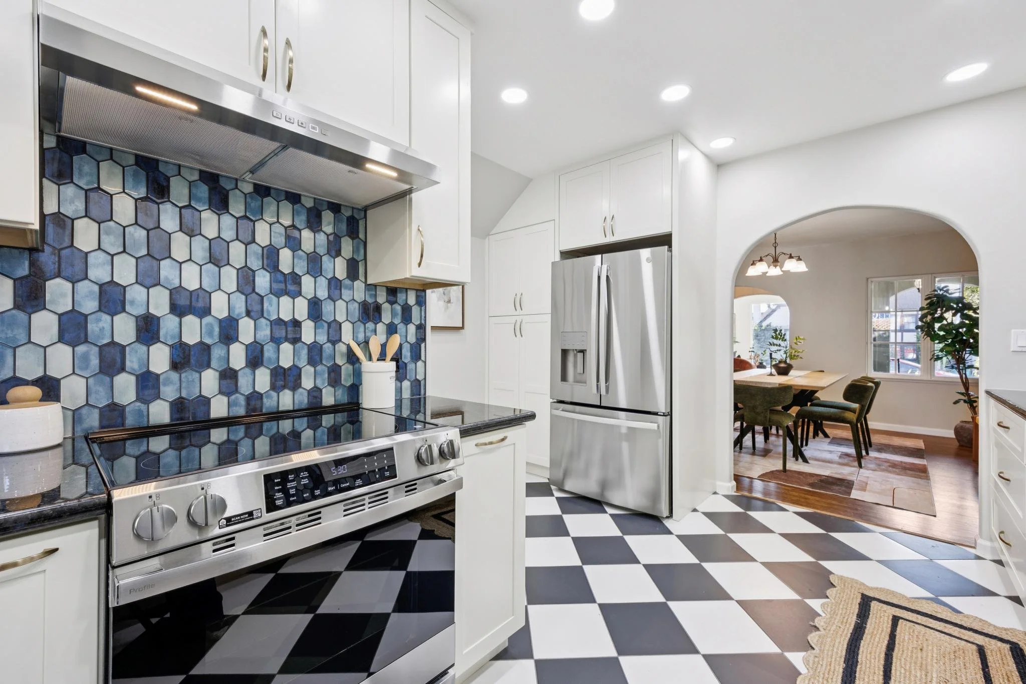 A kitchen with white cabinets, a stainless steel refrigerator, and a stove with a black and white checkered tile floor. There is a blue hexagonal tile backsplash and an arch leading into a dining area with a wooden table, chairs, and a hanging light 