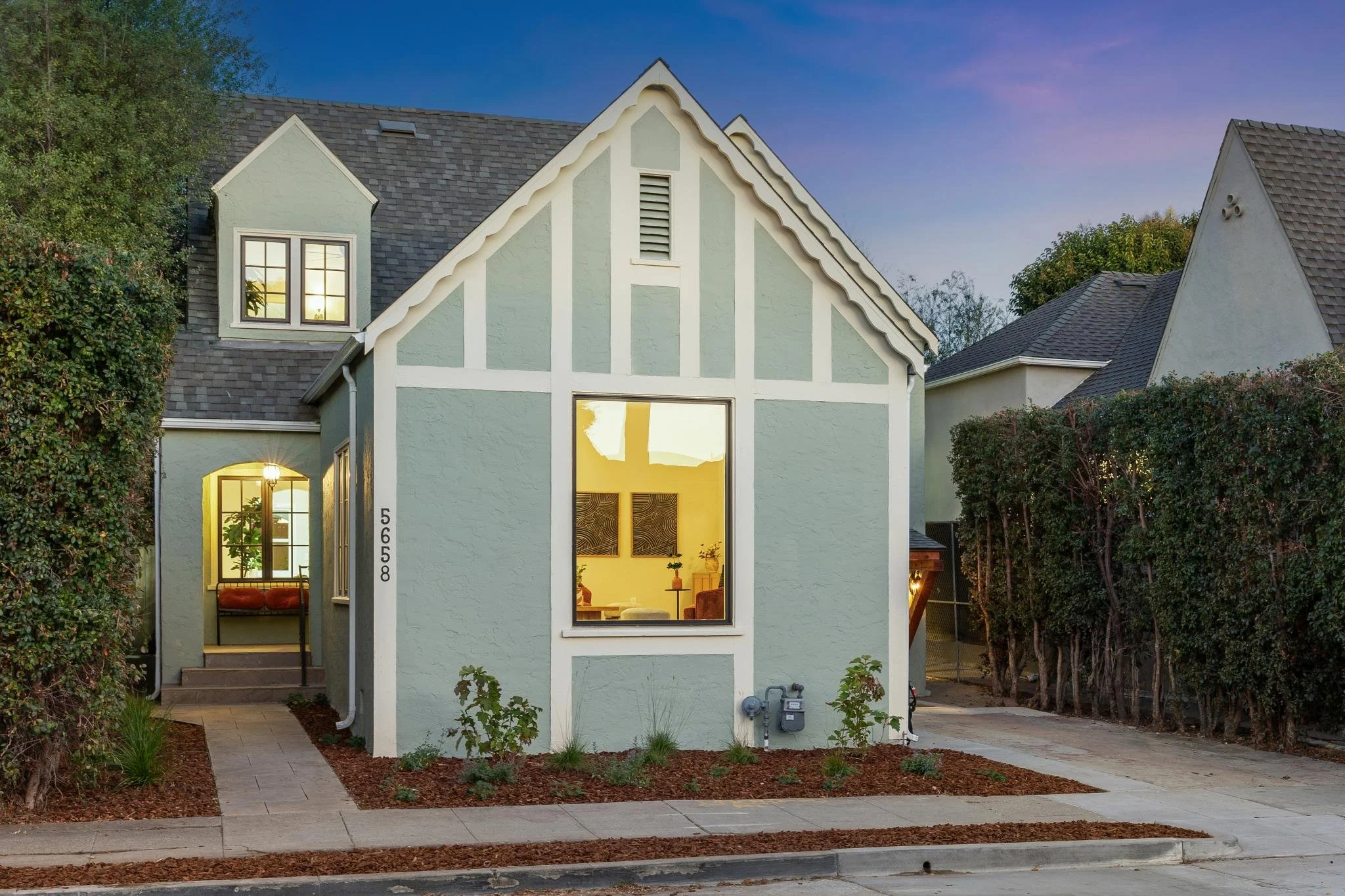 A light green two-story house with a steep gray shingle roof, large front windows, and a small front garden with a sidewalk.