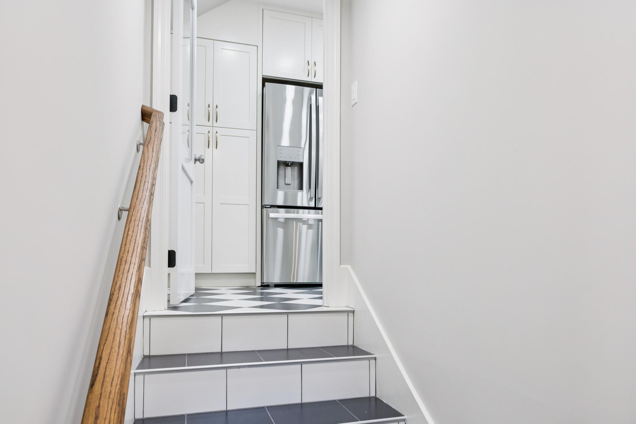 Inside view of a stairway with black and white checkered floor, white walls, a wooden handrail on the left, leading to a small landing with white cabinetry and a stainless steel refrigerator in a kitchen.