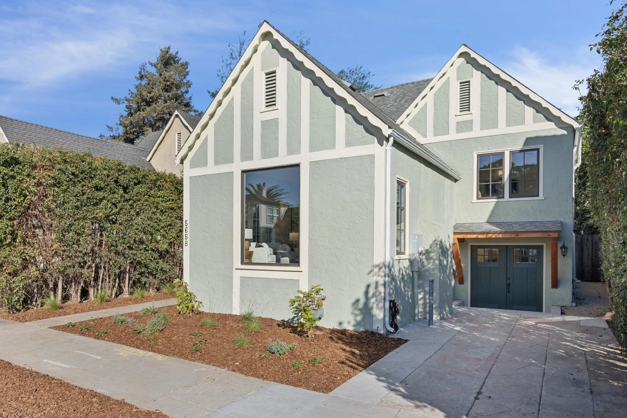 Modern two-story house with a light green exterior, large windows, and a garage on the right side. The front yard has a paved walkway, some small plants, and a mulched garden area. Tall trees and a blue sky are visible in the background.