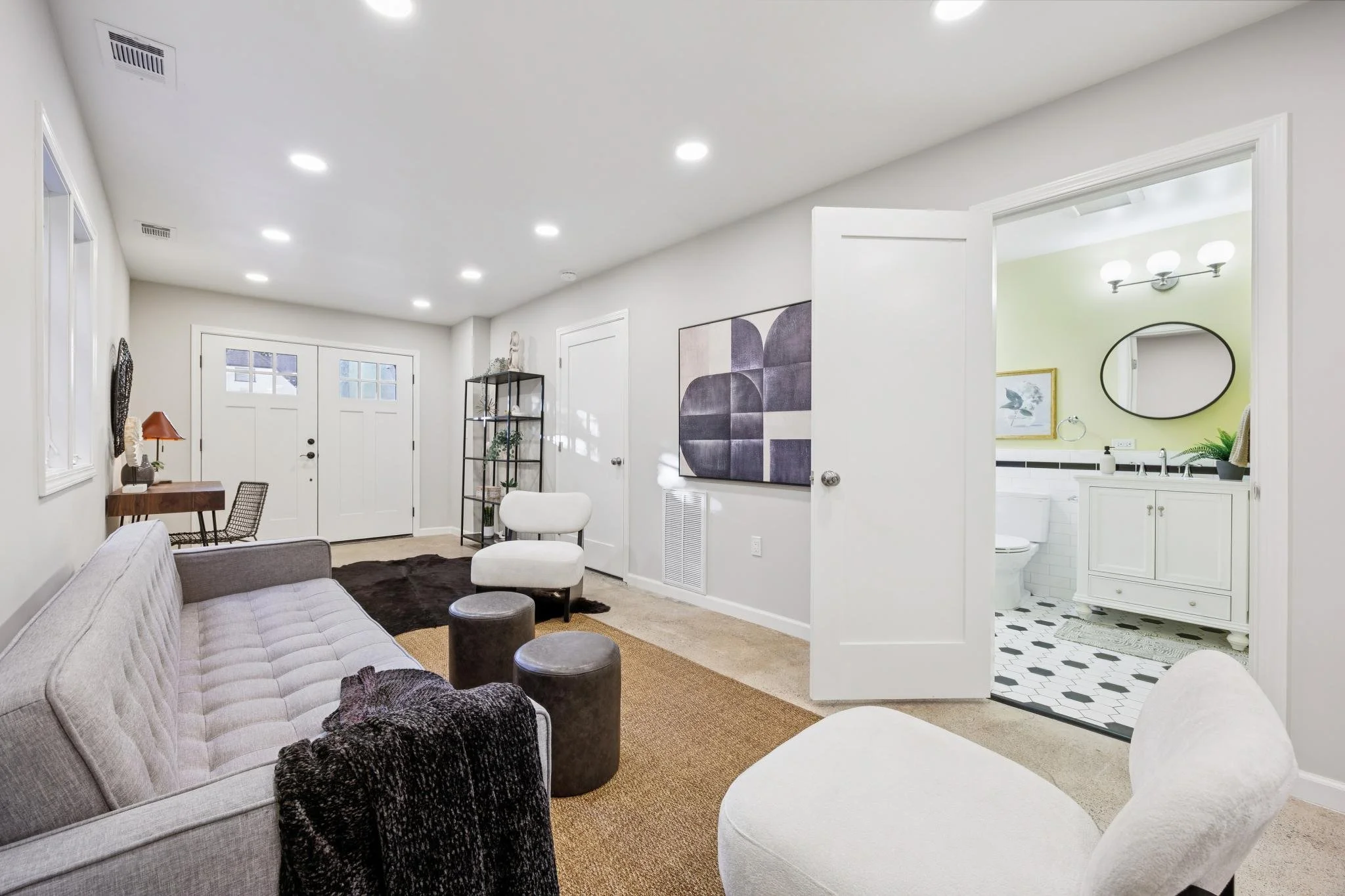Living room with a gray tufted sofa, black ottomans, and beige carpet, adjacent to an open bathroom with black and white tiled floor and a green accent wall.