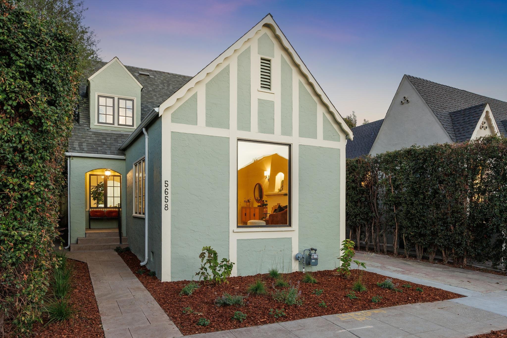 Light blue house with white trim and a large front window, surrounded by a garden with shrubs and small plants, and a paved walkway leading to the entrance, with neighboring houses partially visible.