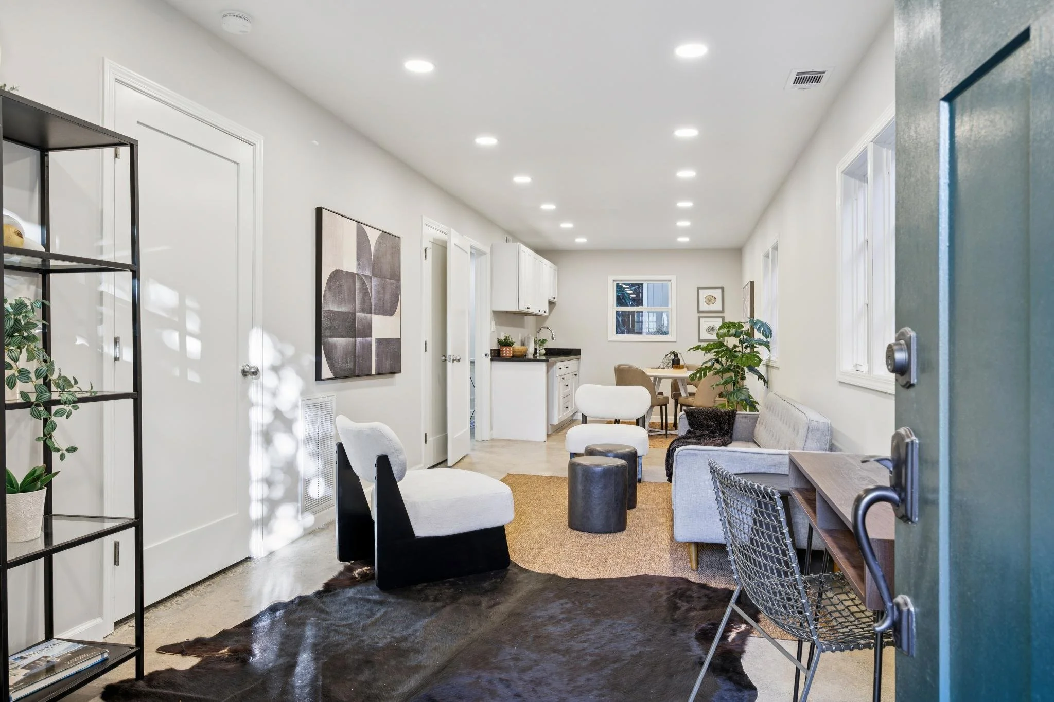 Interior of a modern living space with white walls, multiple large windows, and minimalist furniture including chairs, a sofa, and decorative plants. Recessed ceiling lights illuminate the room.