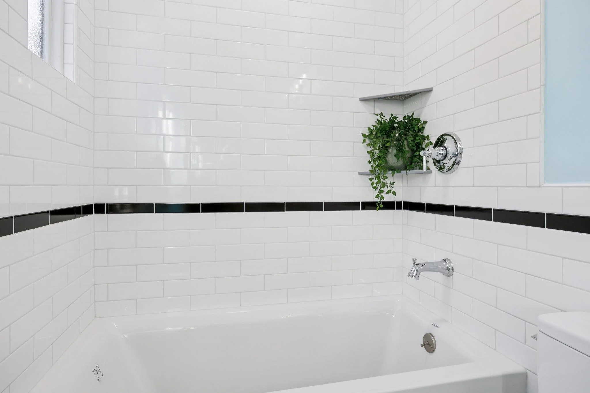 White tiled bathroom with a built-in bathtub, a faucet, a shower controls, a shelf with a green plant, and a small window.