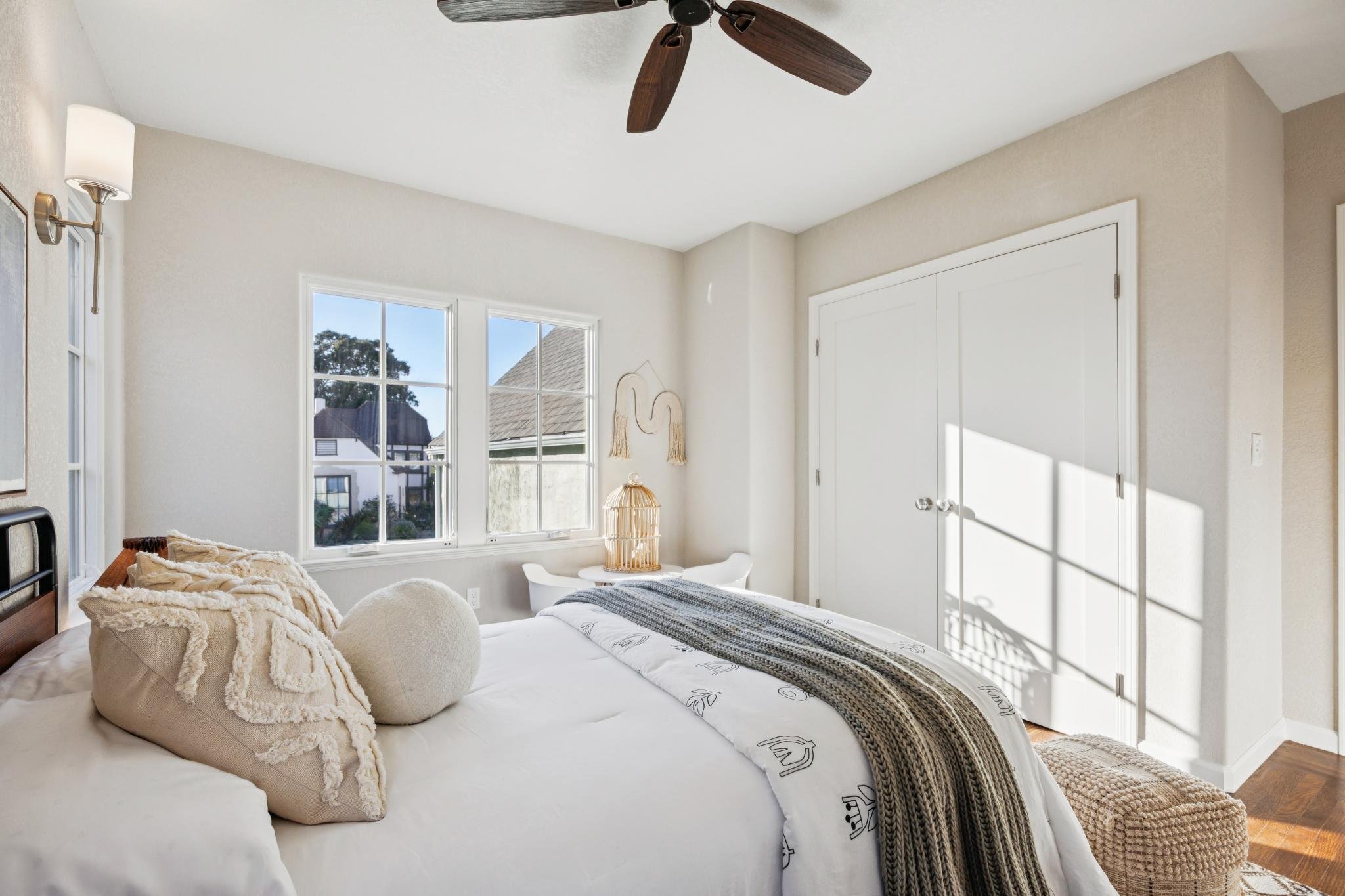 Bright bedroom with beige walls and white trim, two windows with views of neighboring houses and trees, ceiling fan with dark wood blades, wall-mounted light fixture, and a door. A bed with white bedding, a knit blanket, and plush pillows is partiall