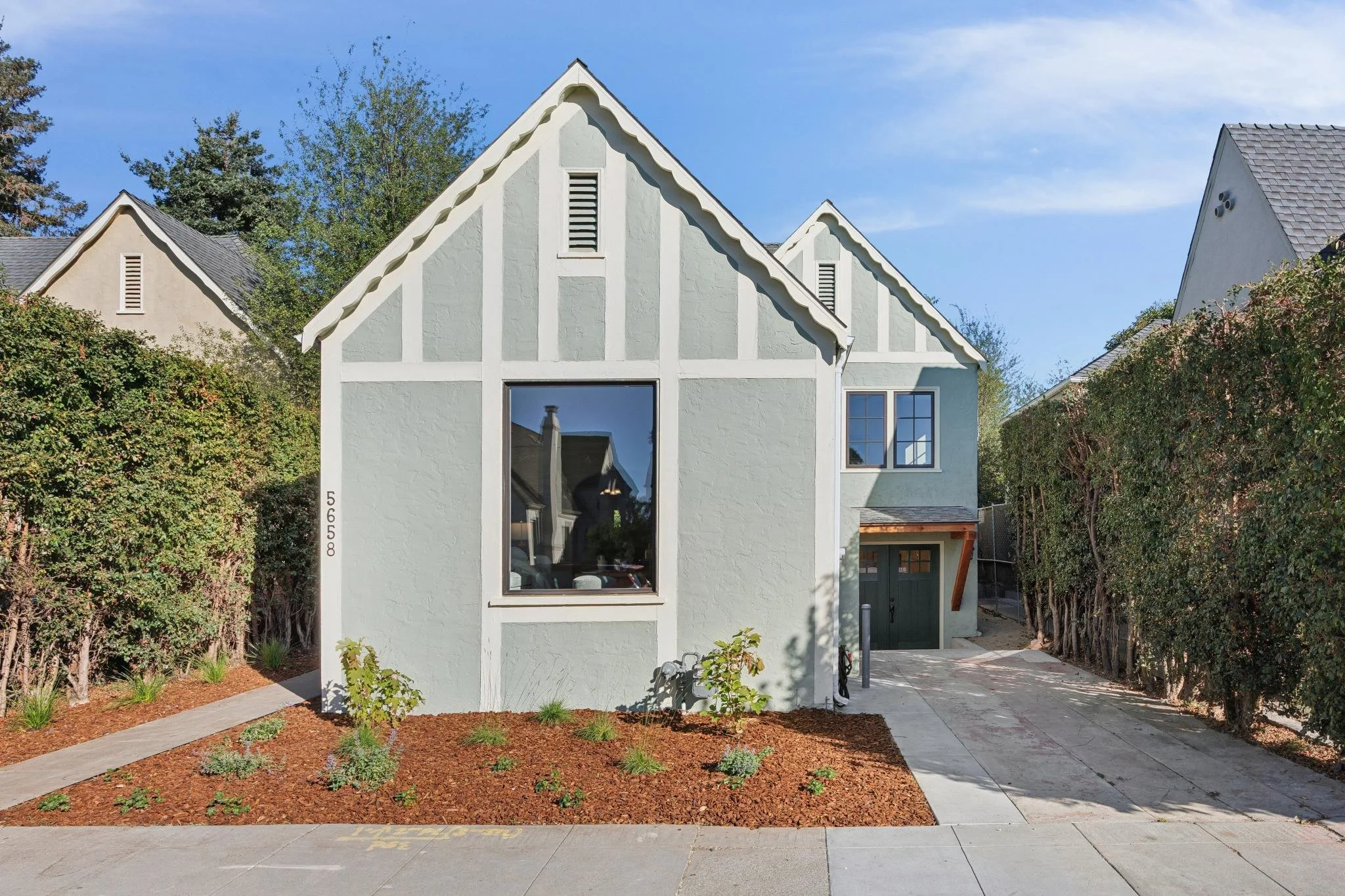 Front view of a modern, light-colored house with a peaked roof, large windows, and a small landscaped garden with mulch and young plants in the front.