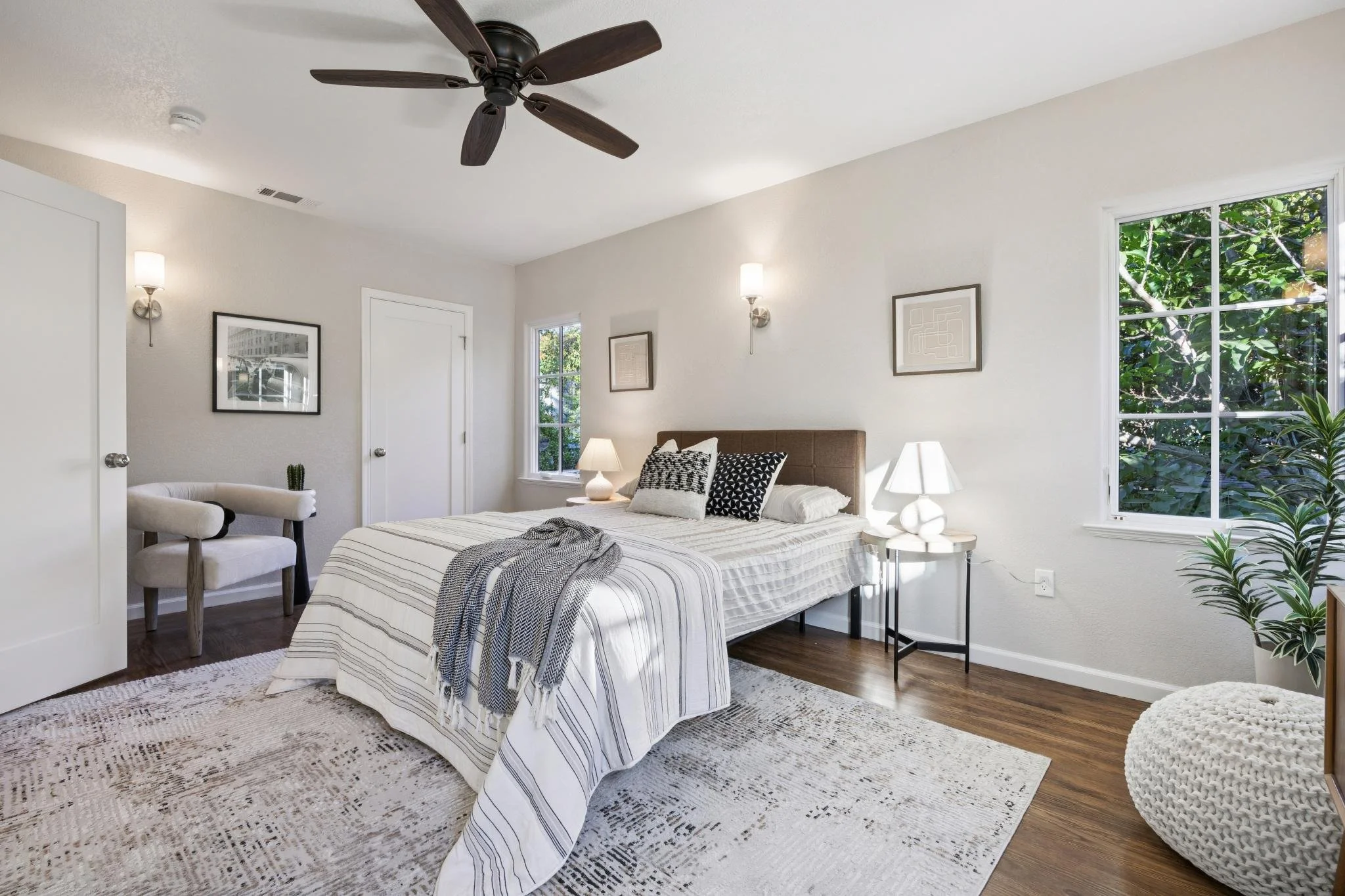 Bright bedroom with white walls, dark wood floors, a bed with striped bedding, nightstands with lamps, a window with green foliage outside, framed artwork on the walls, a ceiling fan, and a large rug.
