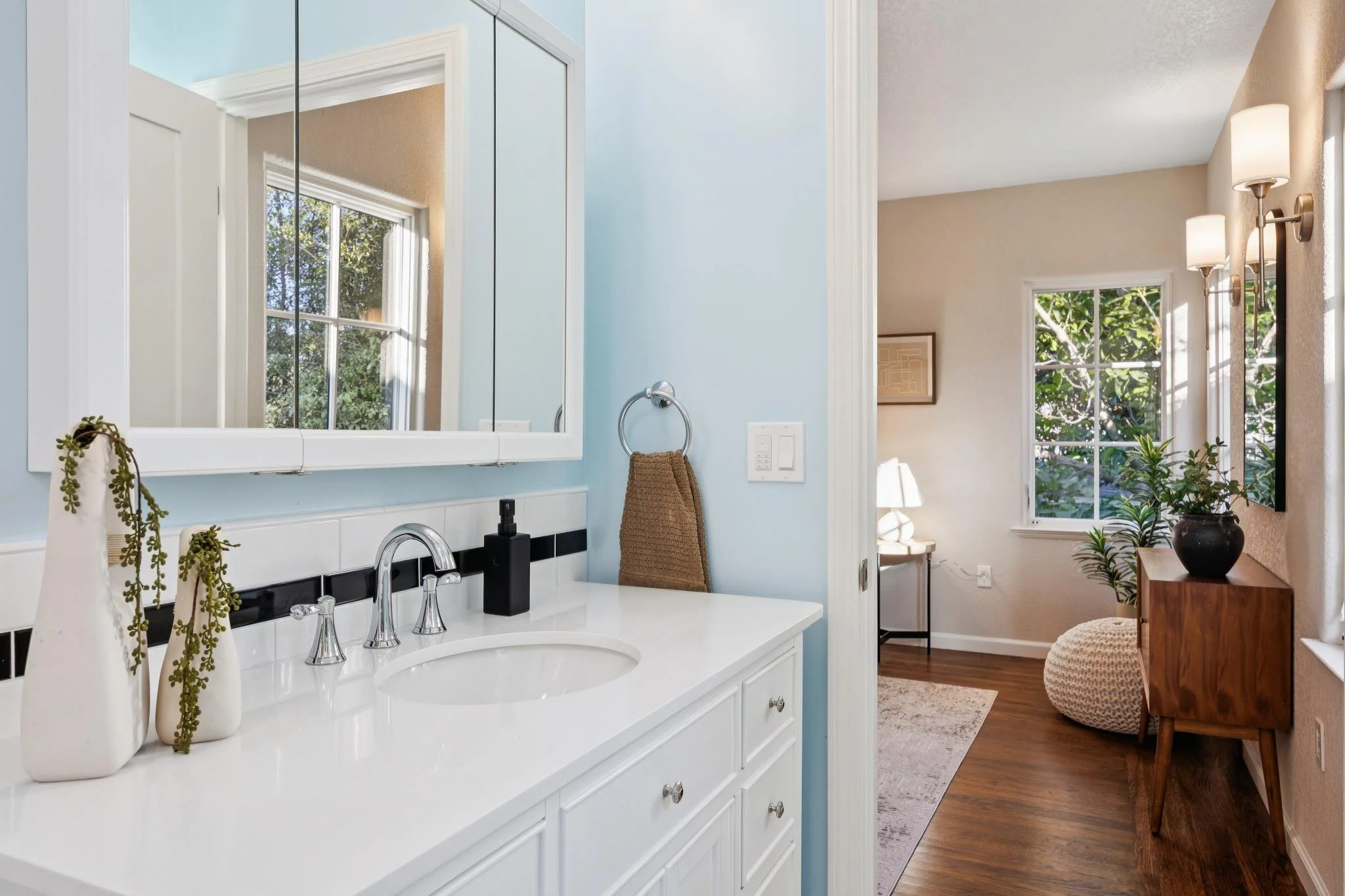 A bathroom vanity with a white countertop, a sink, and a mirror. Decor includes potted plants and a small decorative item. A towel is hanging on a wall-mounted ring. There is a window with a view of trees outside, and part of a living room with large