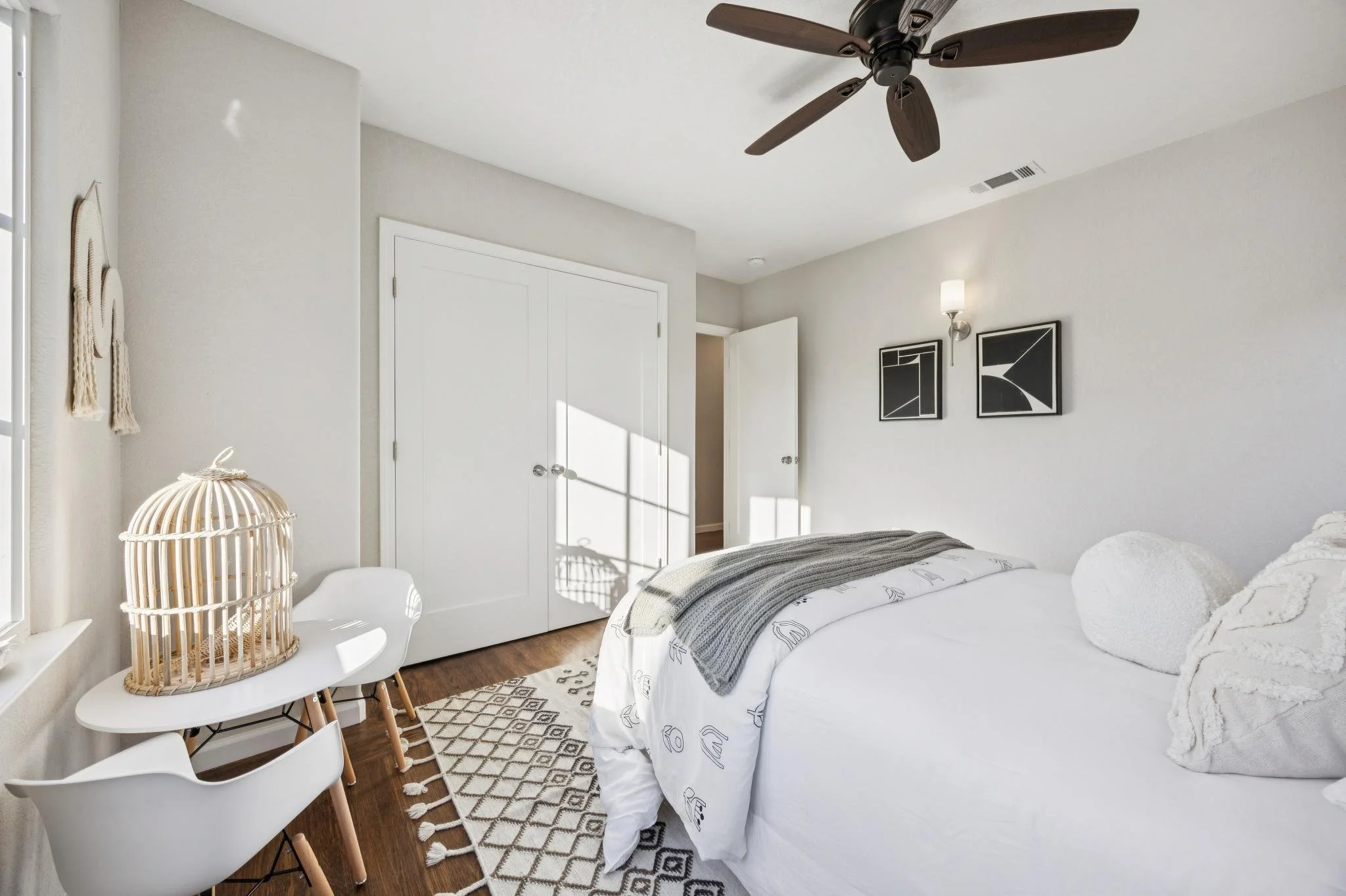 A cozy bedroom with white walls, a bed with white linens and decorative pillows, a ceiling fan, modern black wall art, a small white table with chairs, a woven basket, and a geometric rug on wooden flooring.