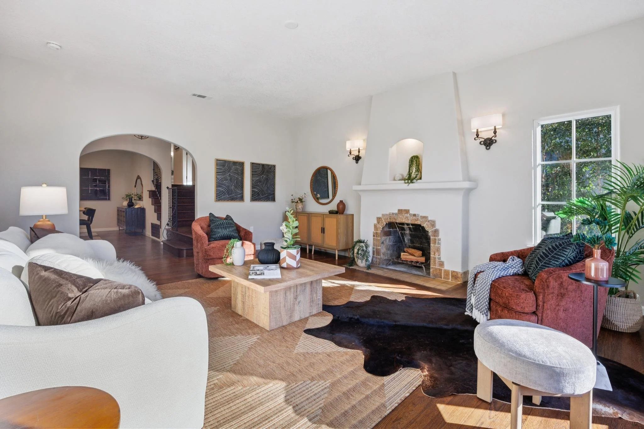 Bright living room with white walls, a fireplace, large window, various sofas, armchairs, wooden coffee table, artwork, plants, and decorative items.