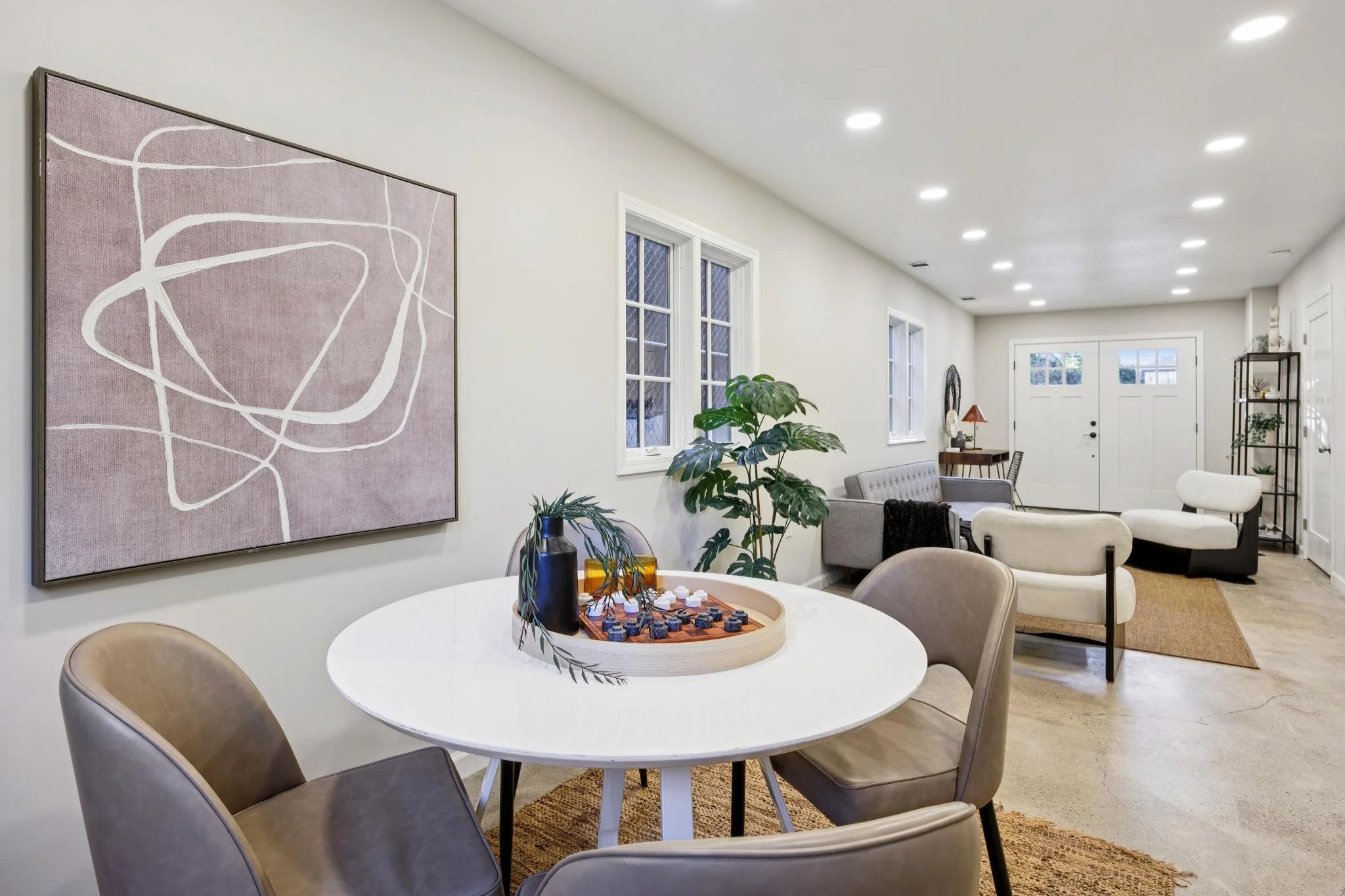 A living room with a white round dining table, tan chairs, a gray sofa, potted plants, and a wall art with abstract pink and white design, illuminated by ceiling lights.