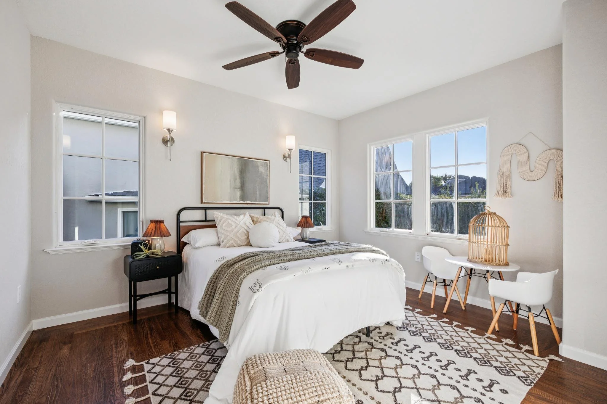 A bedroom with white walls and large windows, a bed with white bedding and decorative pillows, a black bedside table with a lamp, and a small round table with white chairs and a wicker lamp, decorated with wall art and natural decor.