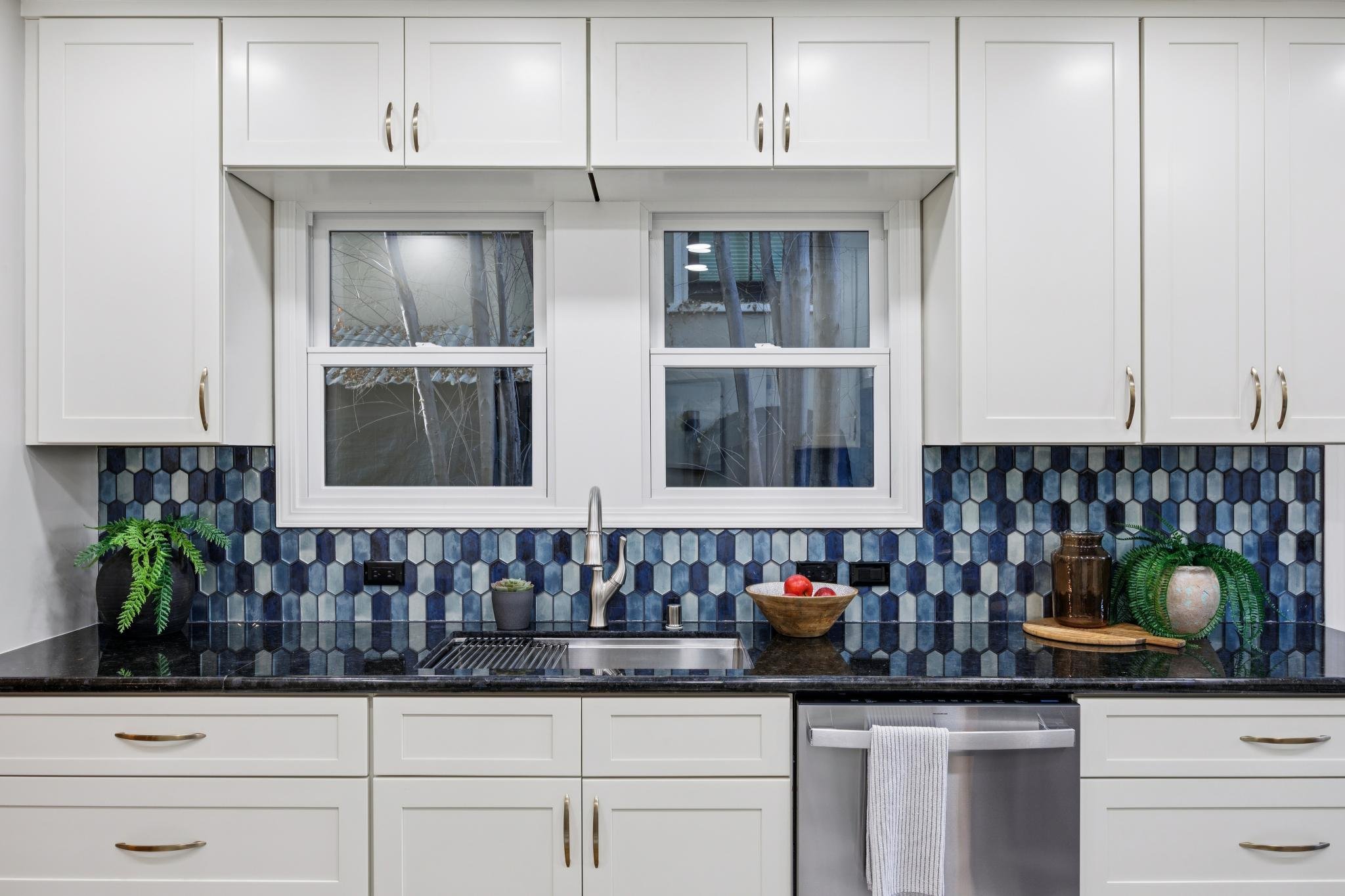A modern kitchen with white cabinets, black countertops, and a blue hexagonal tile backsplash. There are two windows above the sink, with reflections of outdoor trees. Decor items include potted plants, a wooden bowl with apples, and vases.