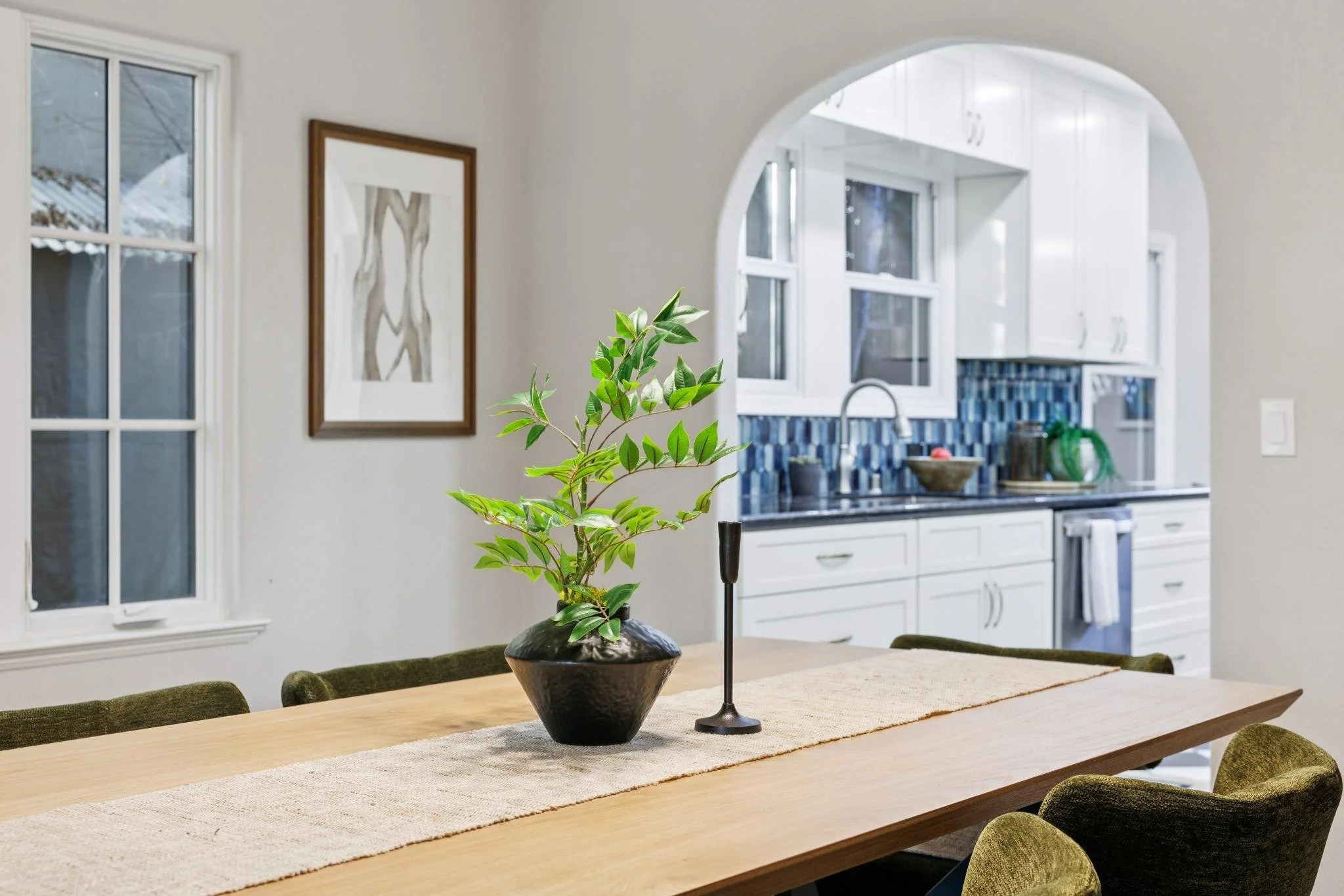 A wooden dining table with a beige table runner, a black ceramic planter with green leafy plant, and a black candle holder, situated in a bright room with white walls, large windows, and a kitchen in the background.