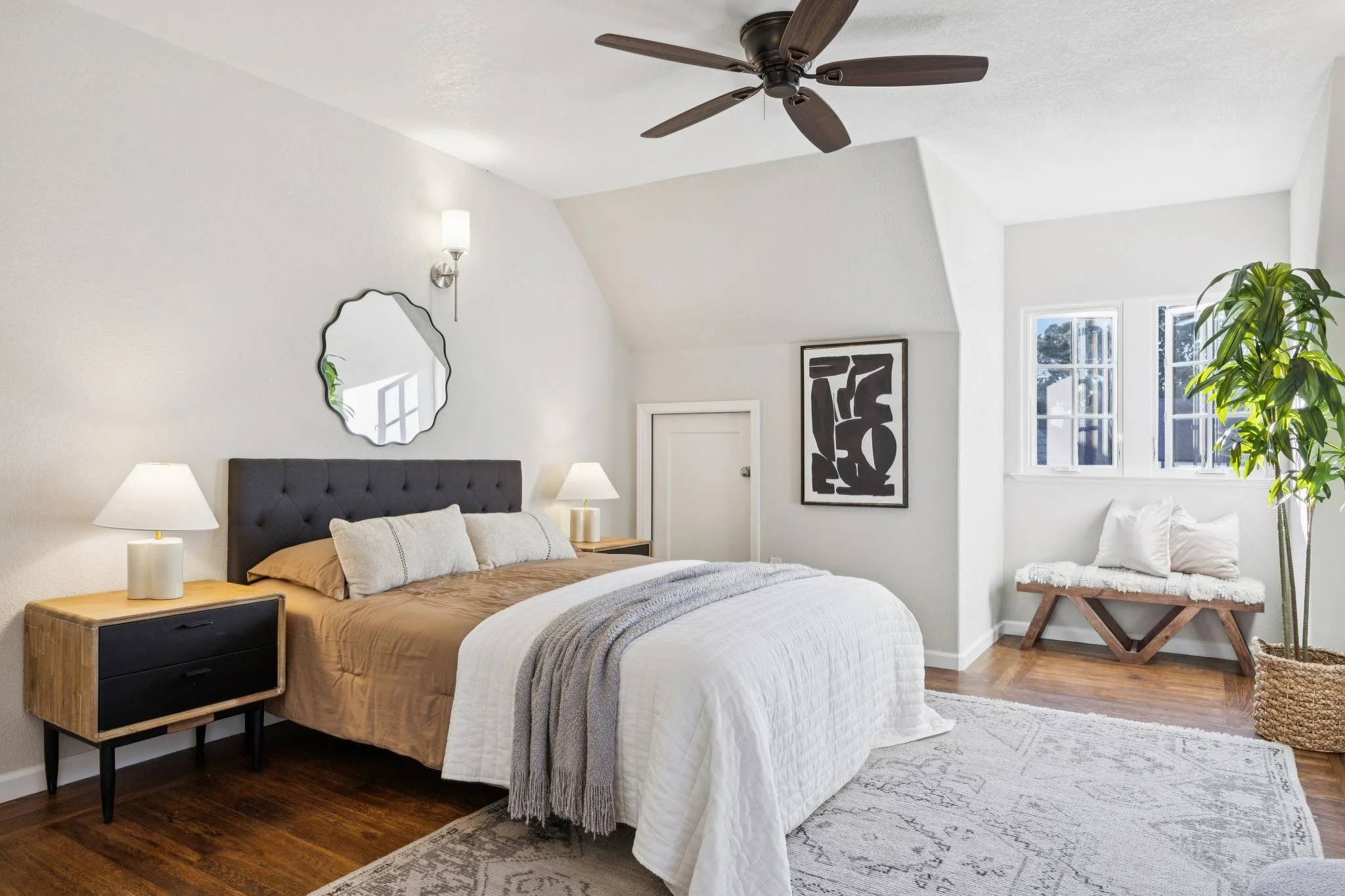 A bedroom with white walls and a sloped ceiling, featuring a bed with brown and white bedding, a black upholstered headboard, two wooden nightstands with lamps, a mirror, an abstract black and white art piece, a large window with trees outside, a pot