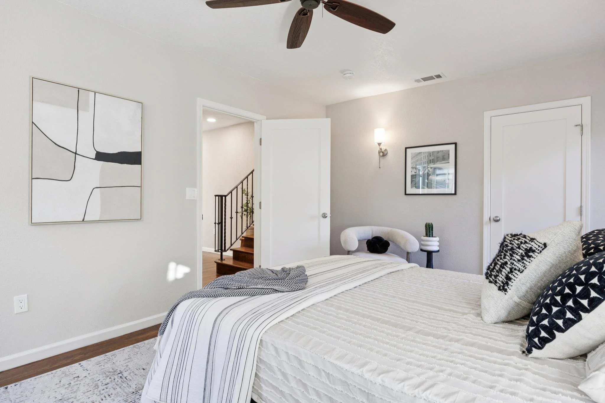 A bedroom with a bed dressed in white and black striped bedding and decorative pillows. There are two contemporary paintings on the walls, a wall-mounted light fixture, a ceiling fan, and a small plant on a side table. An open door reveals a staircas