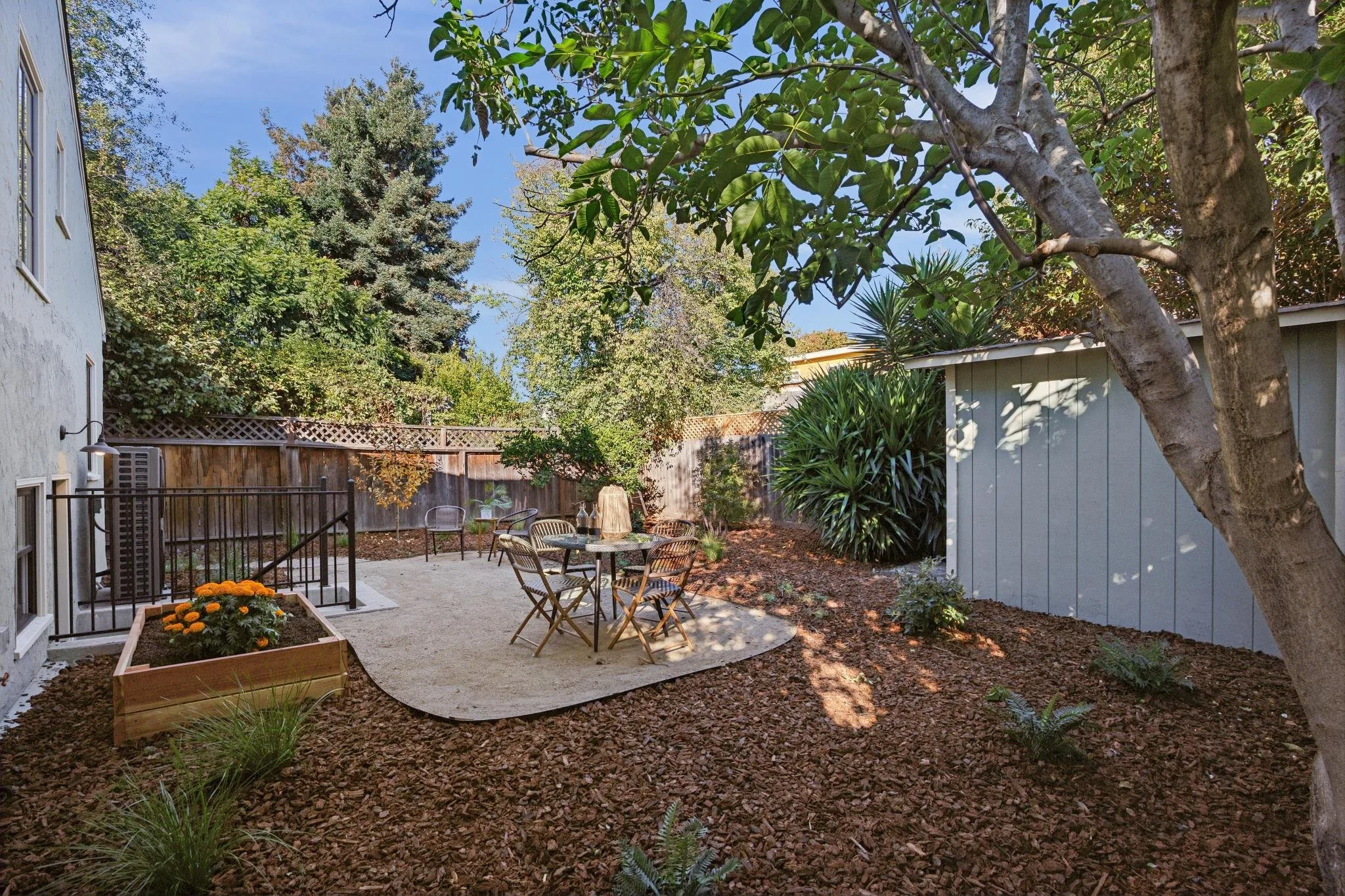 A backyard patio area with outdoor furniture including a round table and chairs, surrounded by trees, plants, and a wooden fence, with a clear blue sky overhead.