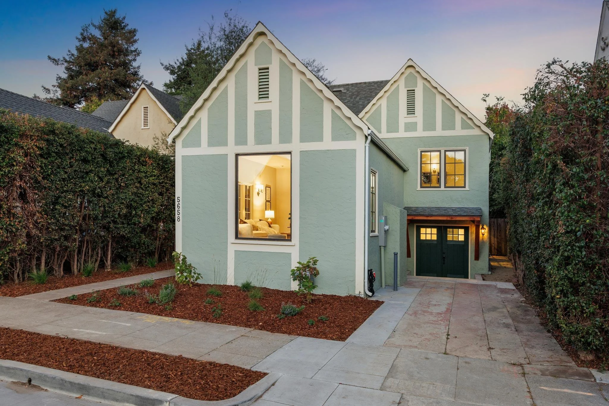A two-story house painted in pastel green with white trim, lit from within, with trees and bushes surrounding it, a sidewalk in front, and a driveway leading to a garage.
