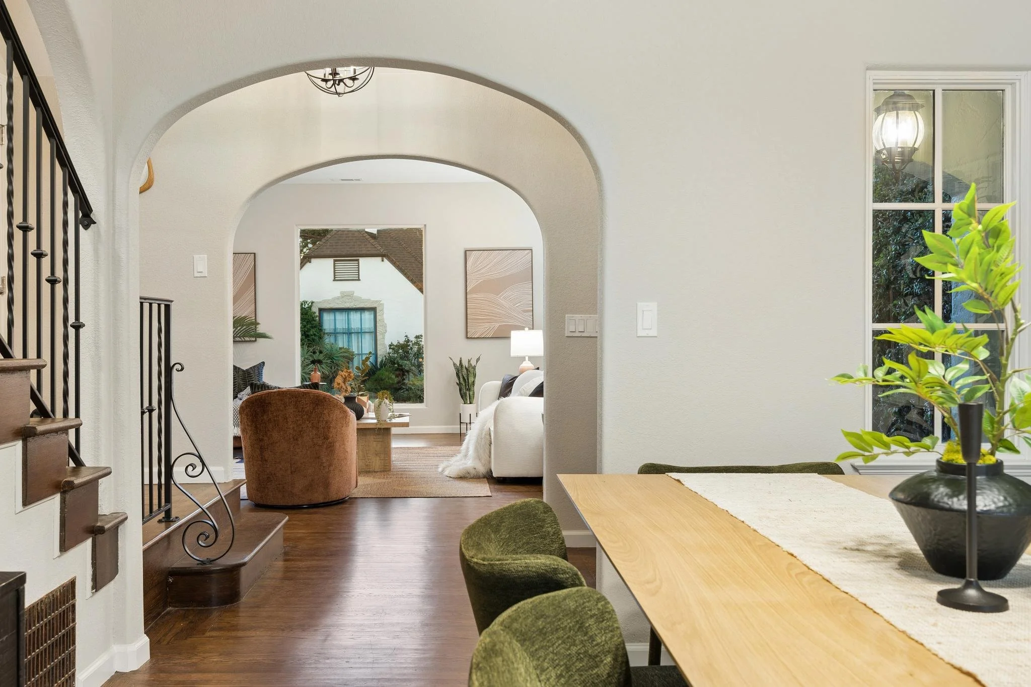 Interior view of a home featuring arched doorways, a dining area with green chairs and a wooden table, and a living room with a white sofa, a brown armchair, and potted plants. Large windows and a ceiling chandelier are visible.