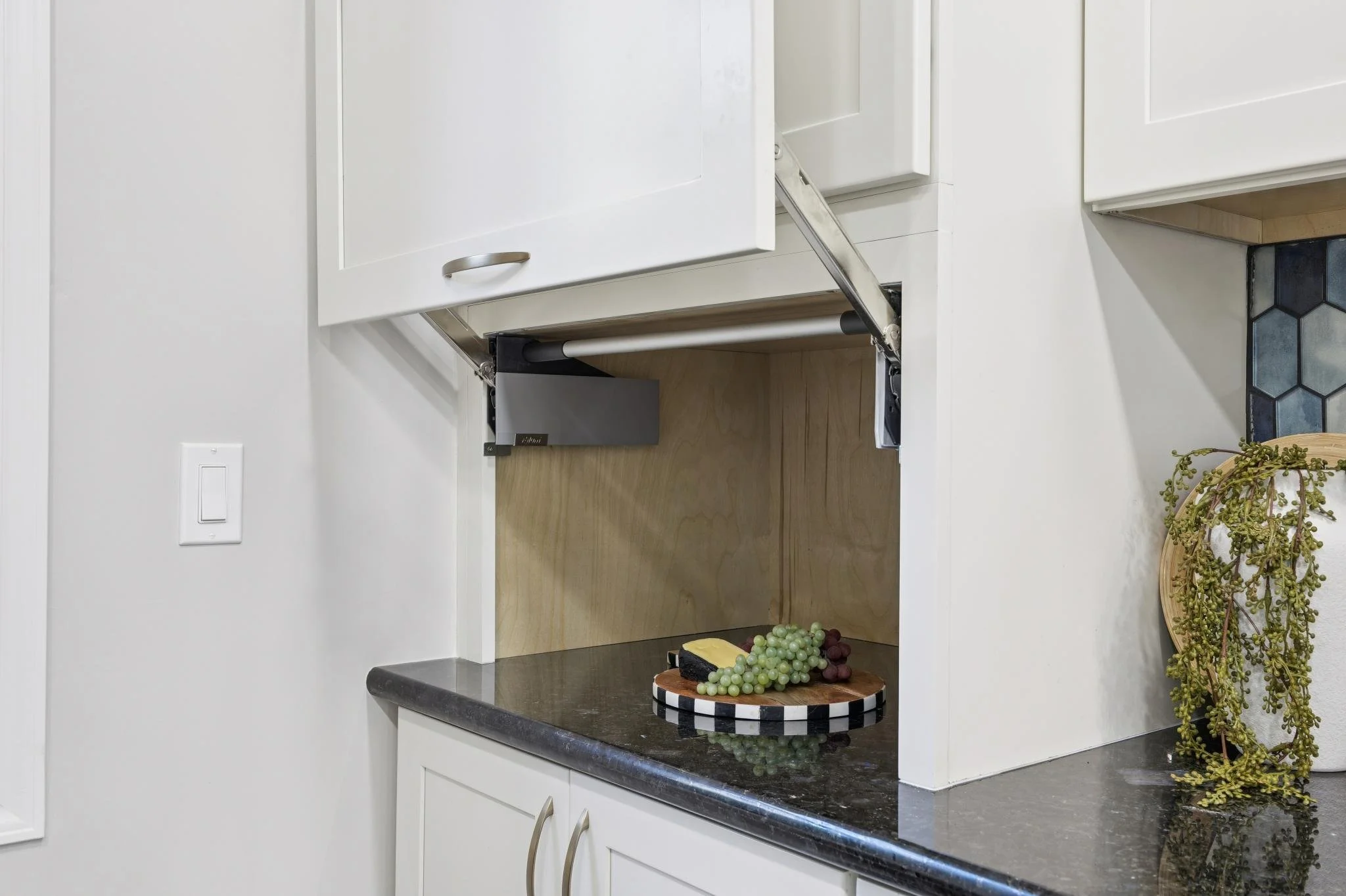 Kitchen with white cabinets, black countertop, and a wooden open shelf. There is a small dish with grapes and cheese on the counter. A basket with decorative plants is on the right side.