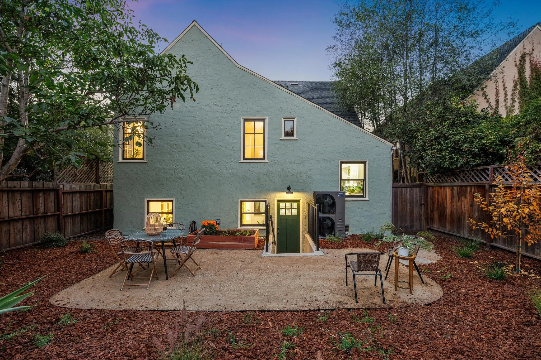 A backyard patio with a green house, some outdoor chairs, a table with flowers, and a fenced yard and trees in the background during early evening.